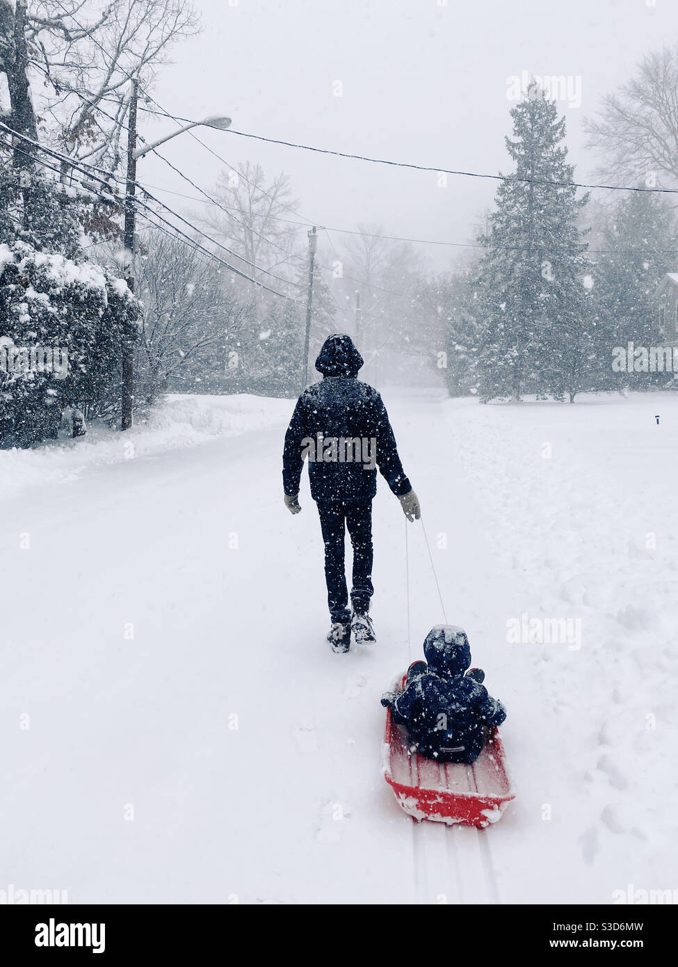 Un père tirant son enfant sur un traîneau dans une rue enneigée dans la banlieue du New Jersey, États-Unis. - Image de stock capturée avec un smartphone
