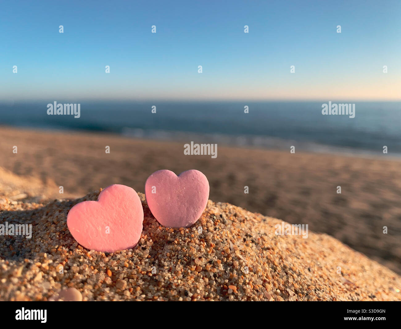 Deux cœurs de bonbons assis sur une plage de sable avec l'océan à l'horizon au coucher du soleil. - Image de stock capturée avec un smartphone