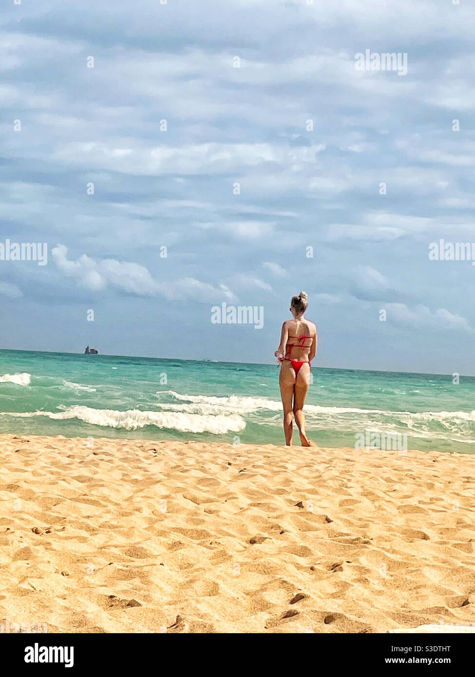 Une femme blonde dans un bikini rouge marche sur le sable vers l'eau turquoise de l'océan avec un bateau de croisière à l'horizon à Miami South Beach, Floride, - Image de stock capturée avec un smartphone