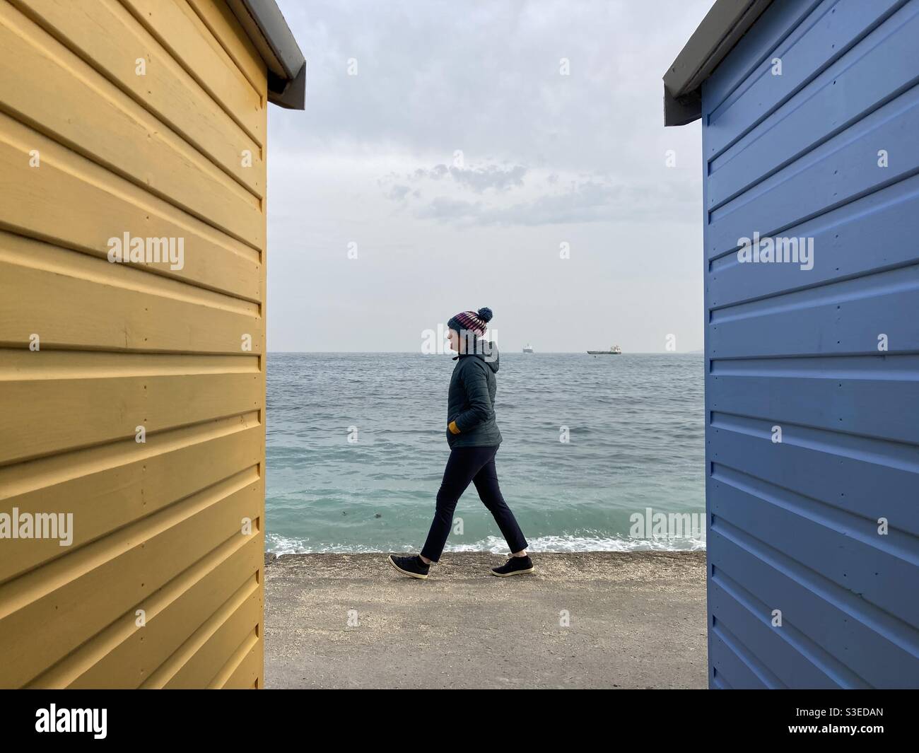 Une femme dans une veste matelassée et un chapeau de laine se promène entre des cabanes de plage à Falmouth, au Royaume-Uni - Image de stock capturée avec un smartphone