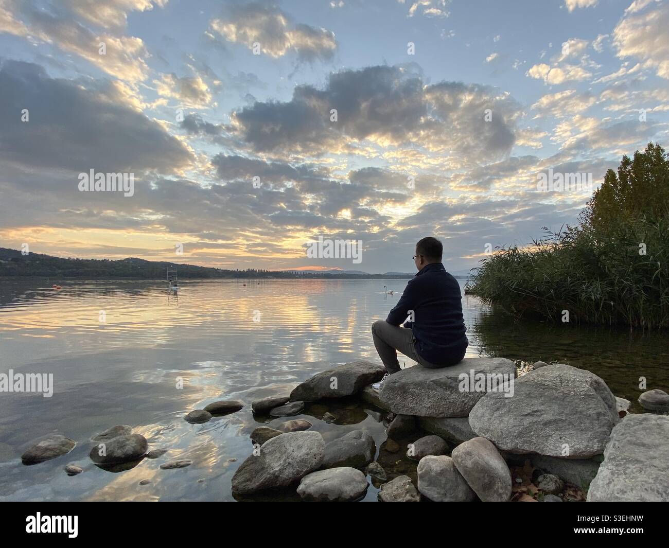 Jeune homme assis au bord du lac de Varese en Italie au coucher du soleil  avec un ciel magnifique et une réflexion Photo Stock - Alamy