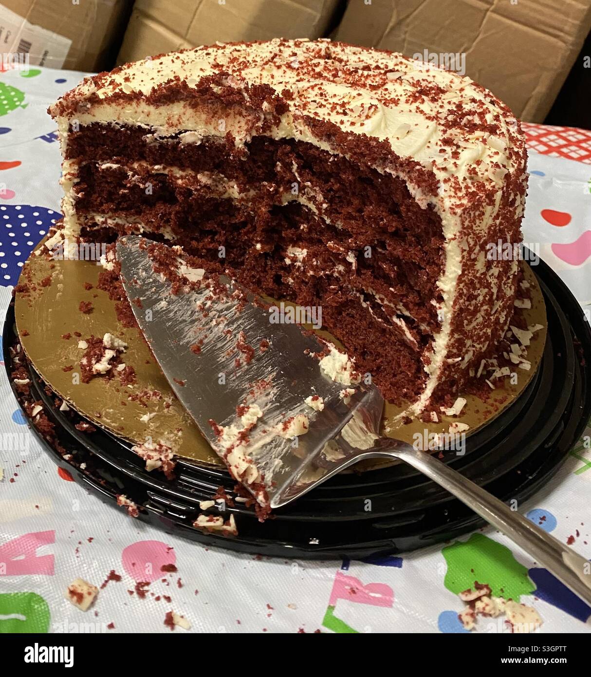 Couper Un Gateau De Velours Rouge Avec Un Coupe Gateau Sur Une Table Recouverte D Une Nappe D Anniversaire En Plastique Et De Chapelure Tout Autour Des Boites En Arriere Plan Montre Que C Est Un Environnement
