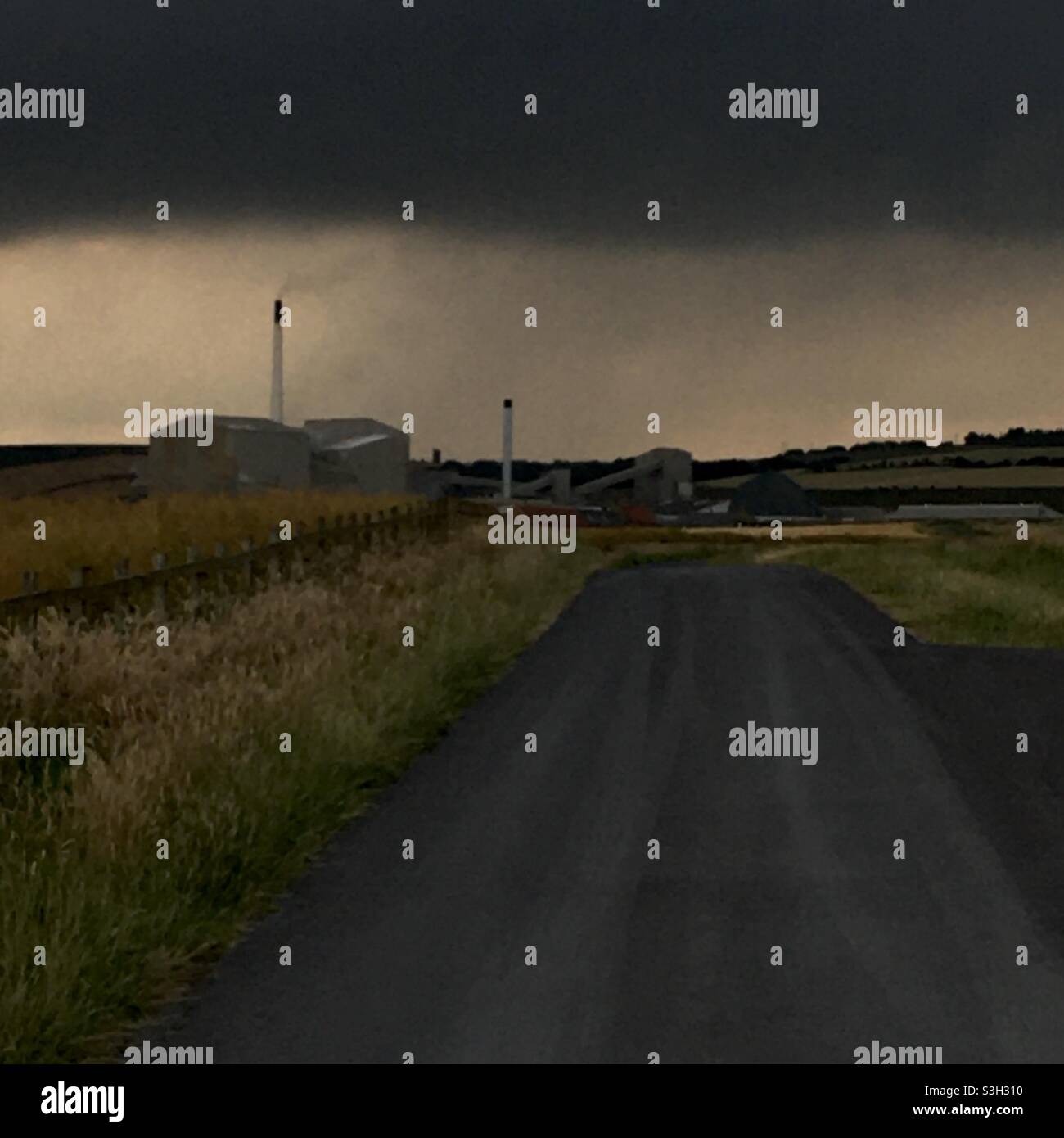 Un paysage agricole industriel minimal sombre avec cheminées d'usine, champs de culture et route et ciel nuageux de pluie sombre Banque D'Images