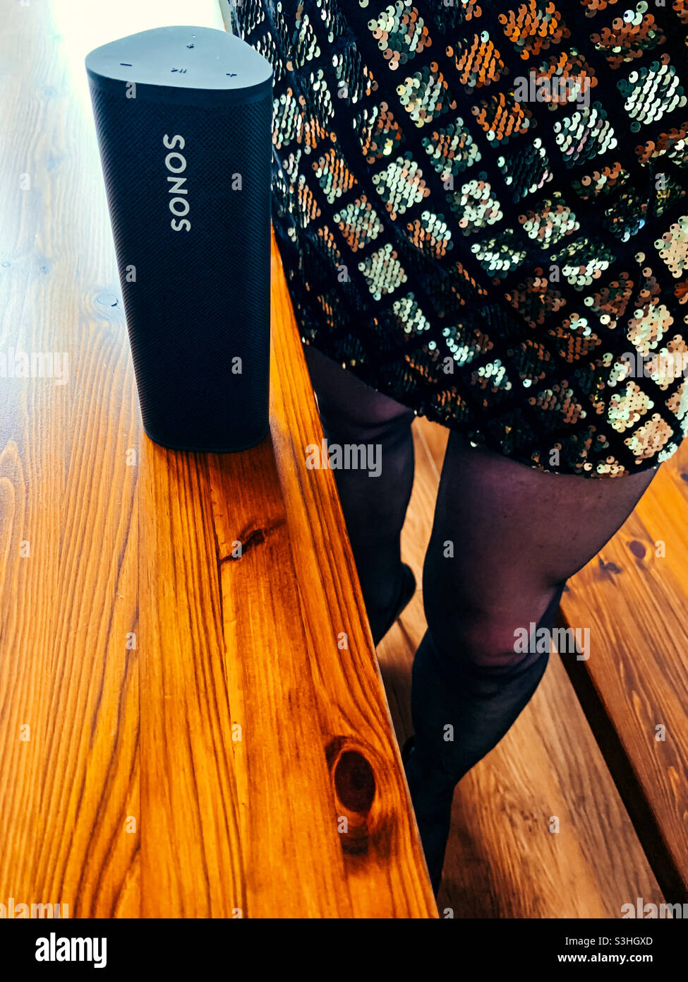 SONOS Roam portable sur une table en bois avec une femme debout à côté d'elle portant une robe en sequins Banque D'Images