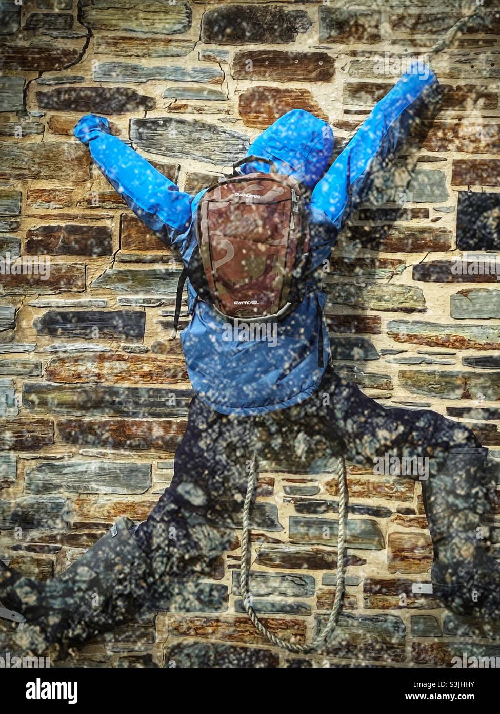 Personne portant un anorak à capuche, un pantalon imperméable et un sac à dos grimpant un mur de pierre - Image de stock capturée avec un smartphone