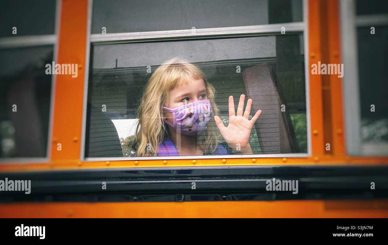 Blonde cheveux fille d'école avec le masque agitant de la fenêtre de bus Banque D'Images