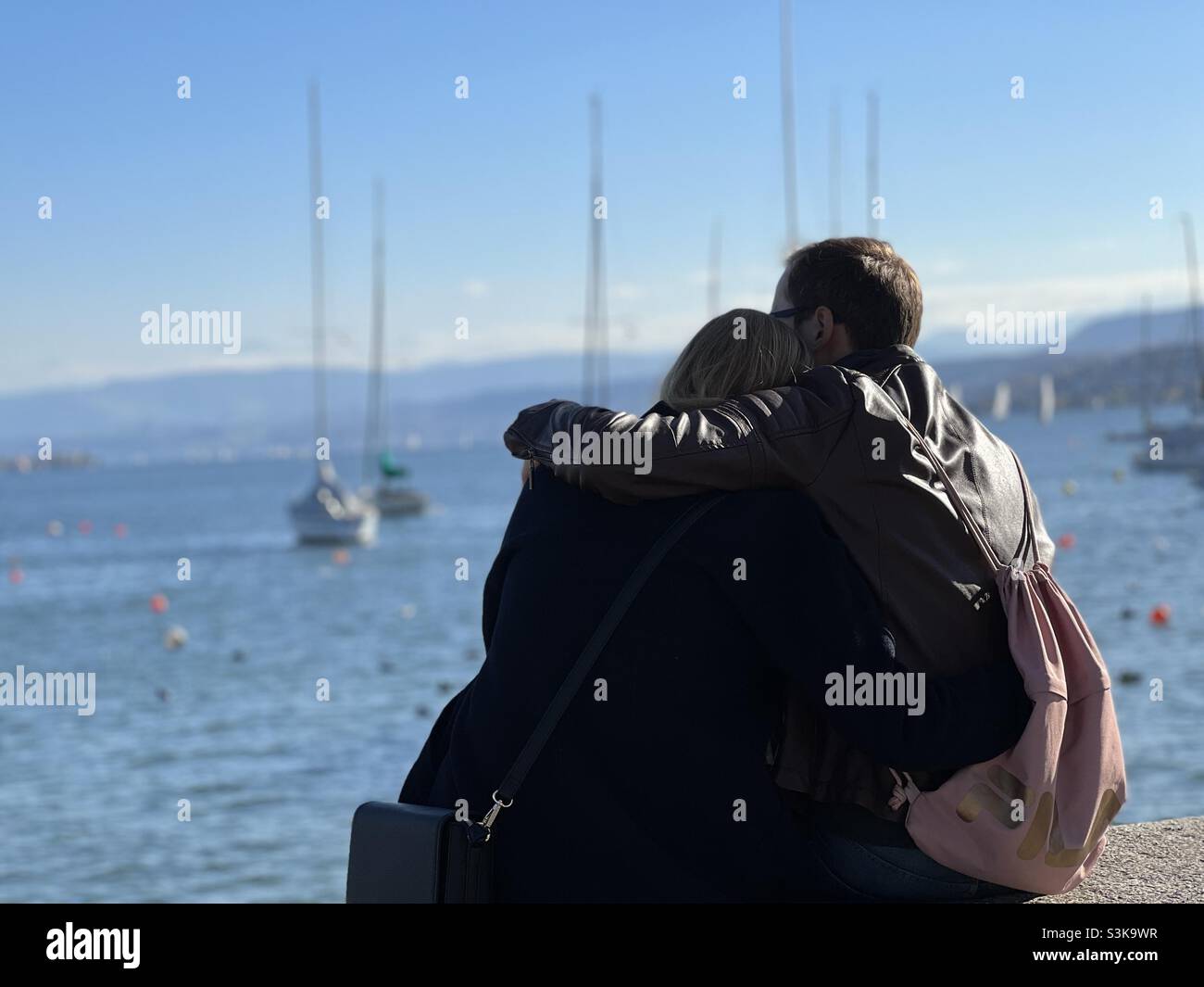 Les jeunes couples hétérosexuels se cuddling ensemble dans la vue arrière.Ils sont assis sur une barrière observant le lac de Zurich, en Suisse. Banque D'Images