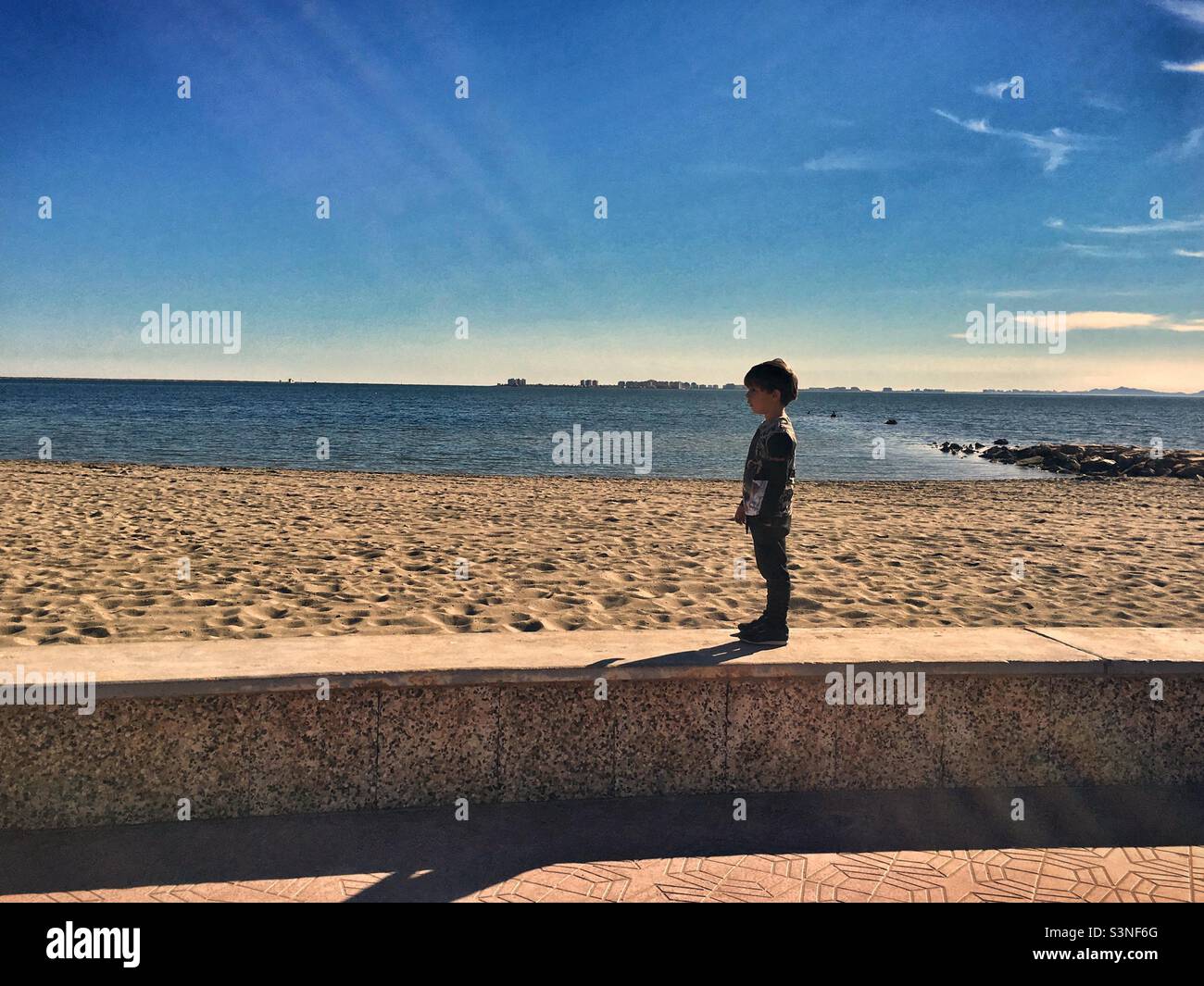 Un petit garçon face aux rayons du soleil sur une plage vide Banque D'Images