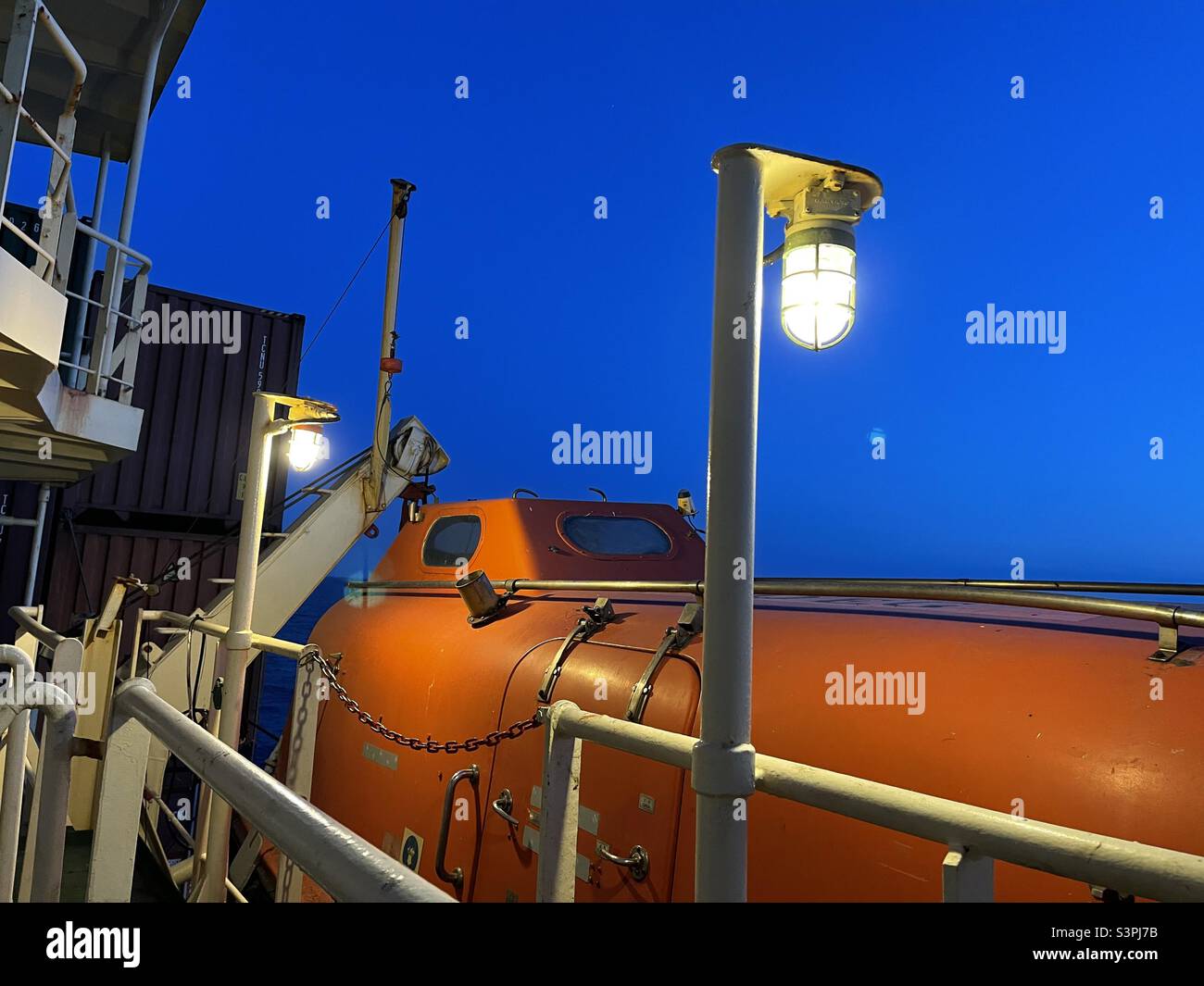 Canot de sauvetage orange fixé côté port du conteneur prêt pour l'urgence éclairé par deux lumières électriques pendant le coucher du soleil . Le bateau navigue en mer Méditerranée pendant le calme du printemps. Banque D'Images