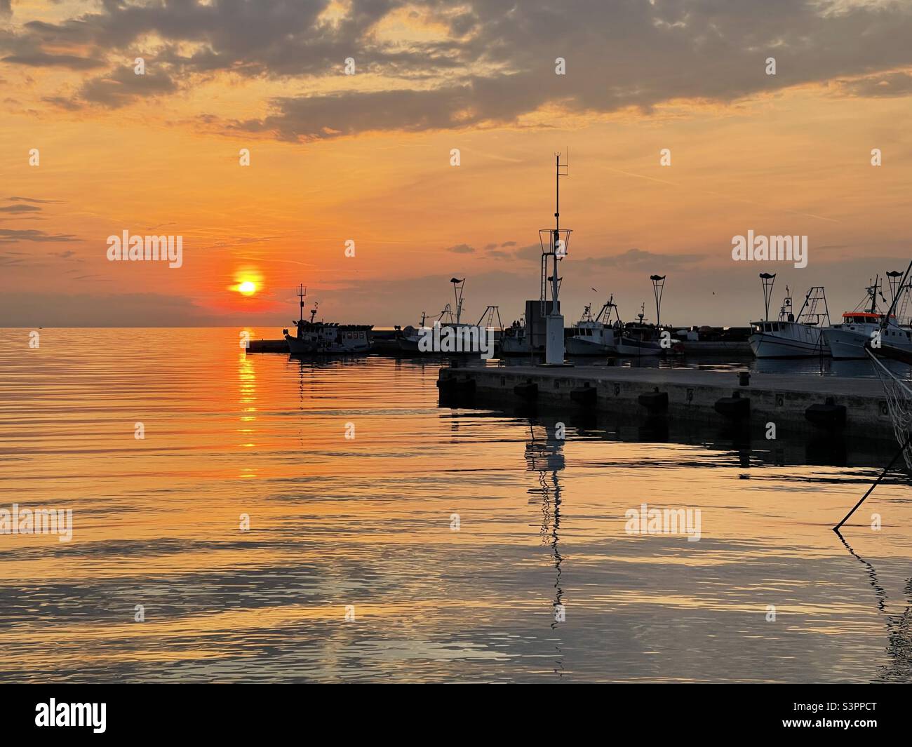 Jetée et port de pêche avec bateaux et petits bateaux pendant le coucher du soleil dans la ville de Koper, Slovénie pendant la saison de printemps et temps calme. À l'horizon est le soleil dans le ciel orange et les nuages gris. Banque D'Images