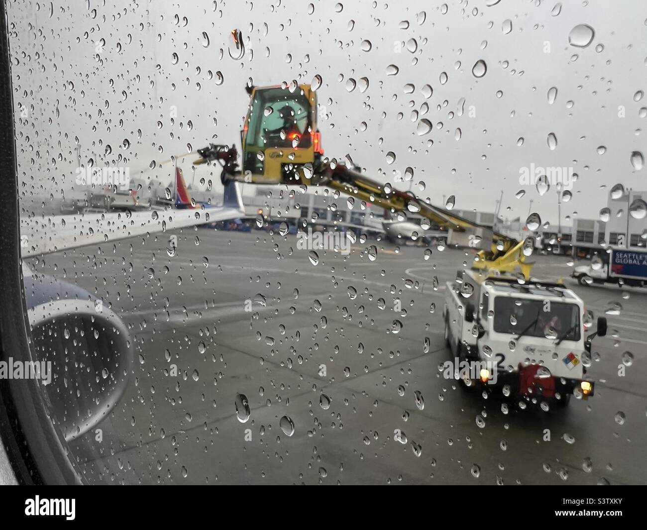 Concentrez-vous sur les gouttes de pluie sur la fenêtre de l'avion passager alors que les travailleurs, en mode adouci, pulvérisent du liquide de dégivrage sur les ailes à l'aide d'un équipement spécialisé en arrière-plan Banque D'Images