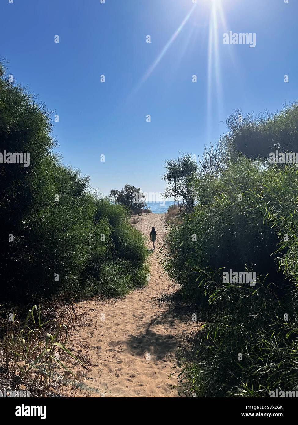 Enfant marchant sur un chemin de sable vers la plage, éclairé par le soleil dans un ciel bleu Banque D'Images