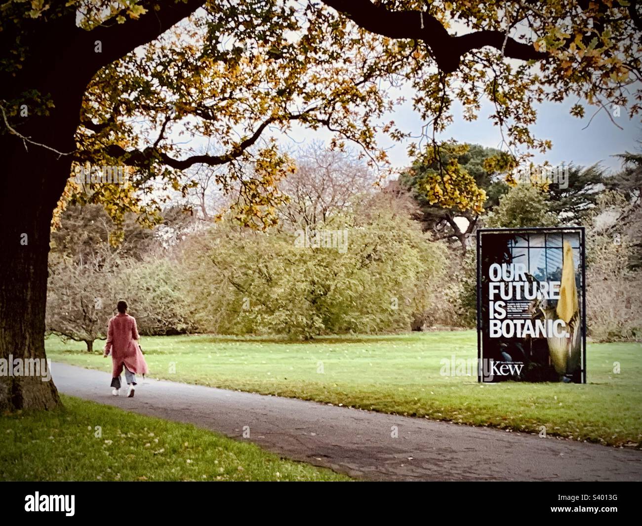Dame portant un long manteau rose traversant les jardins botaniques royaux de Kew avec un panneau indiquant « notre avenir est botanique » Banque D'Images