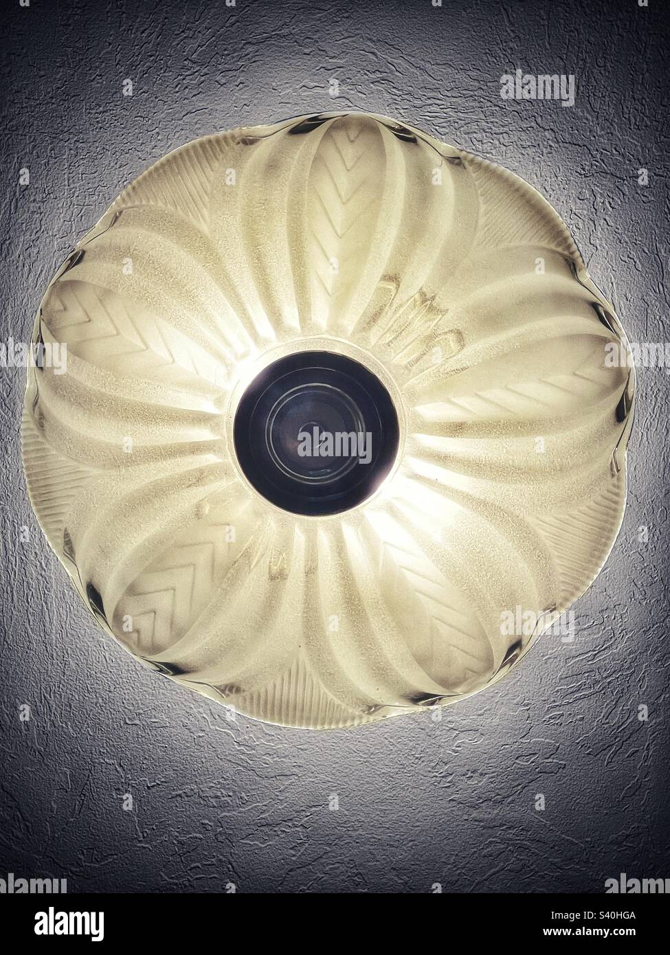 Vue du dessous d'un plafonnier. La lumière est allumée, la lueur éclairant non seulement la gravure sur l'abat-jour en verre, mais aussi la texture sur le plafond. Banque D'Images