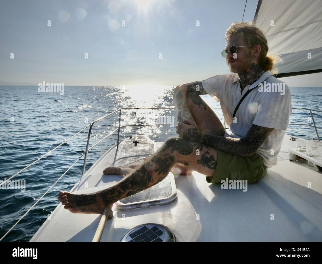 Un homme aux tatouages bénéficie du soleil depuis le pont d'un catamaran sur une mer calme Banque D'Images
