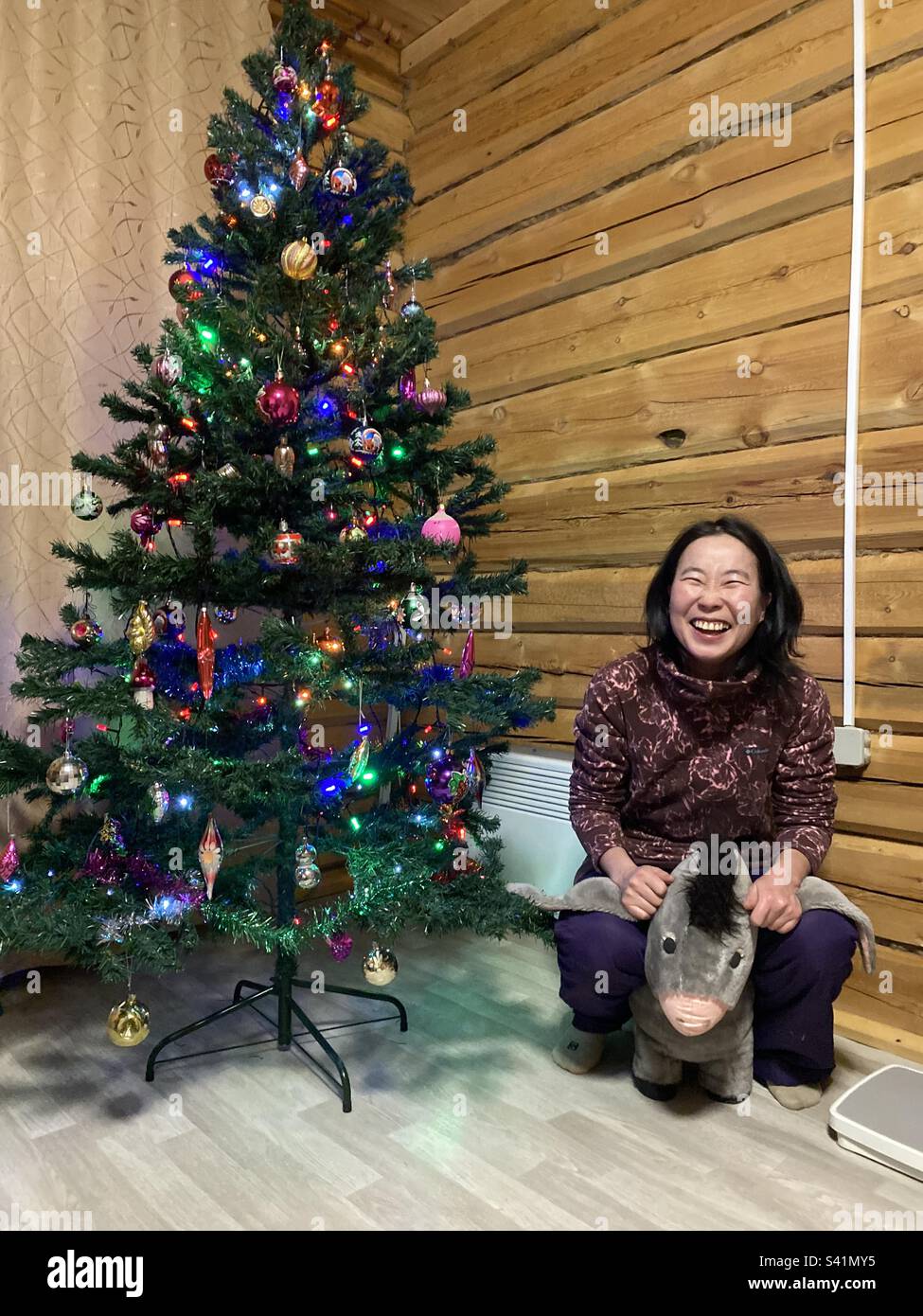 Une fille Yakut rit en s'asseyant avec un âne en peluche, levant ses oreilles sur un arbre de Noël décoré. - Image de stock capturée avec un smartphone