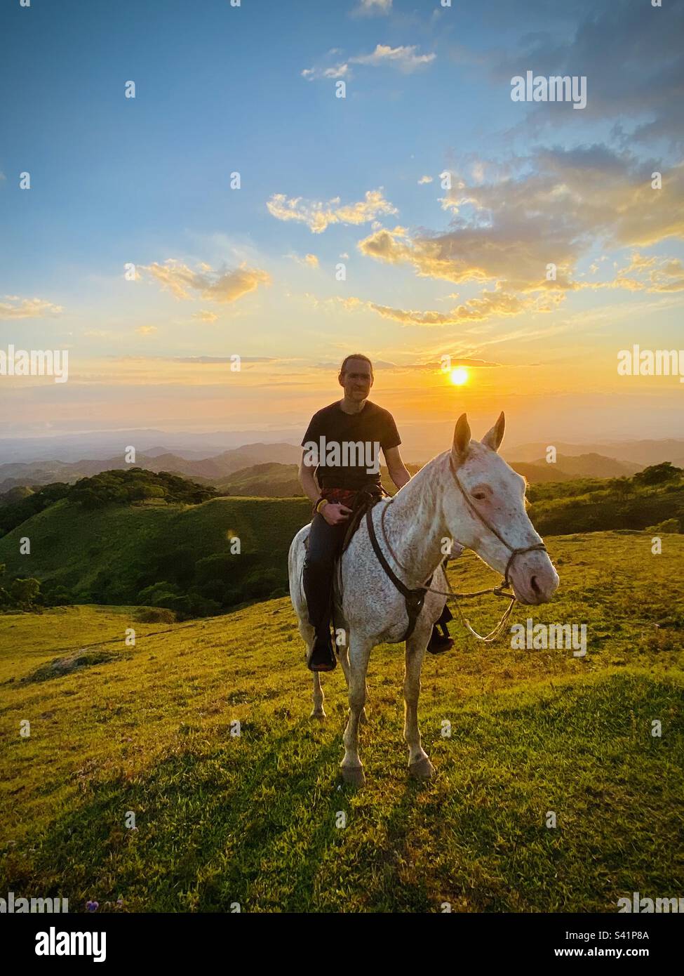 Un homme sur un cheval blanc pose pour une photo tandis que le soleil se couche sur les collines de Monteverde au Costa Rica Banque D'Images