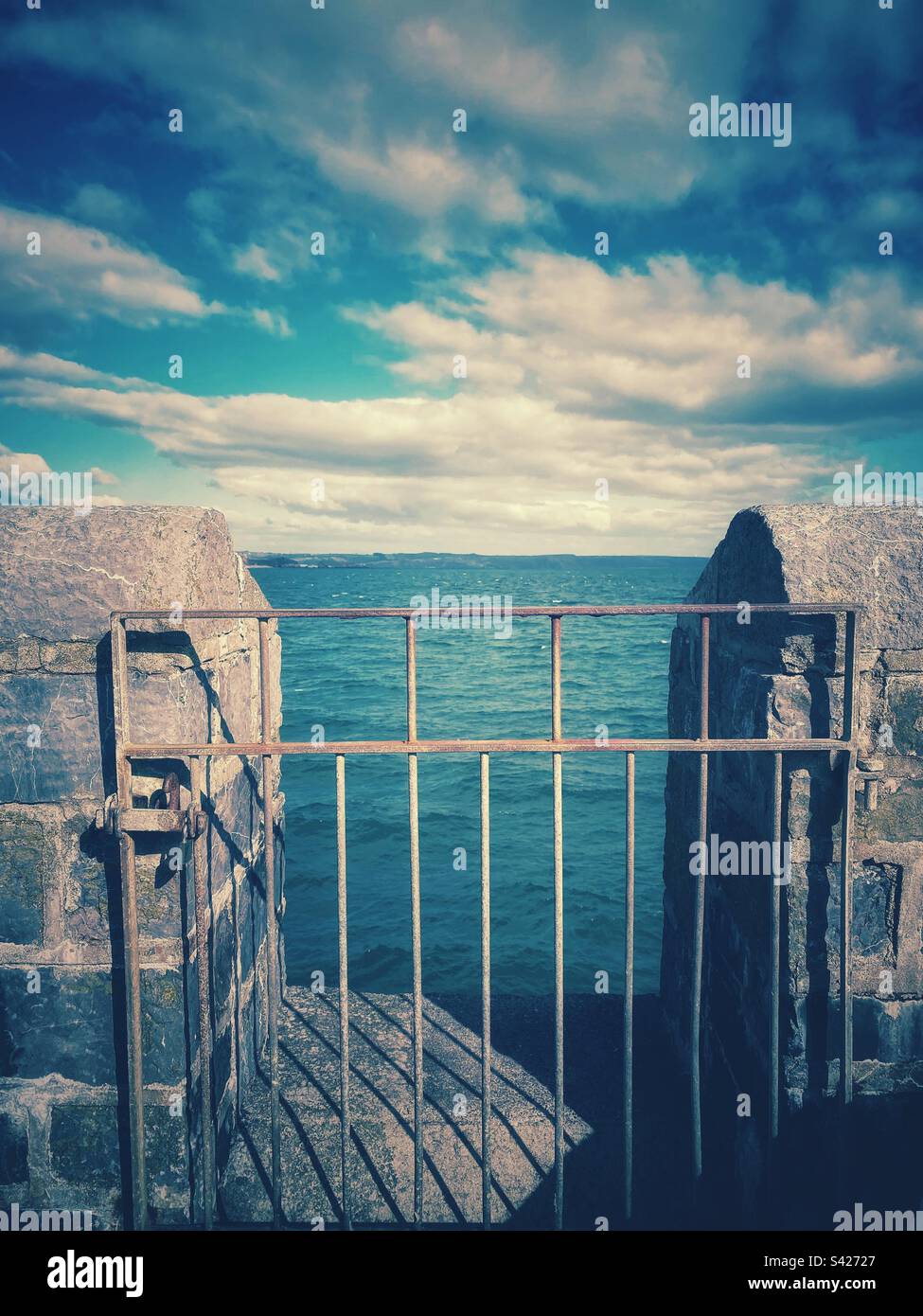 Photographie d'une porte métallique menant à l'océan à travers un vieux mur en pierre. Vue sur la côte Banque D'Images