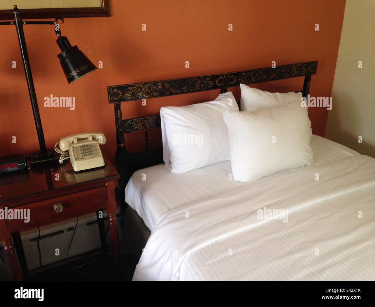 Chambre d'hôtel dans le Vermont avec draps blancs et mode ancien, tête de lit noire ornée avec de beaux détails floraux. Téléphone de style ancien avec boutons-poussoirs. Aussi, mur d'accent orange. Banque D'Images