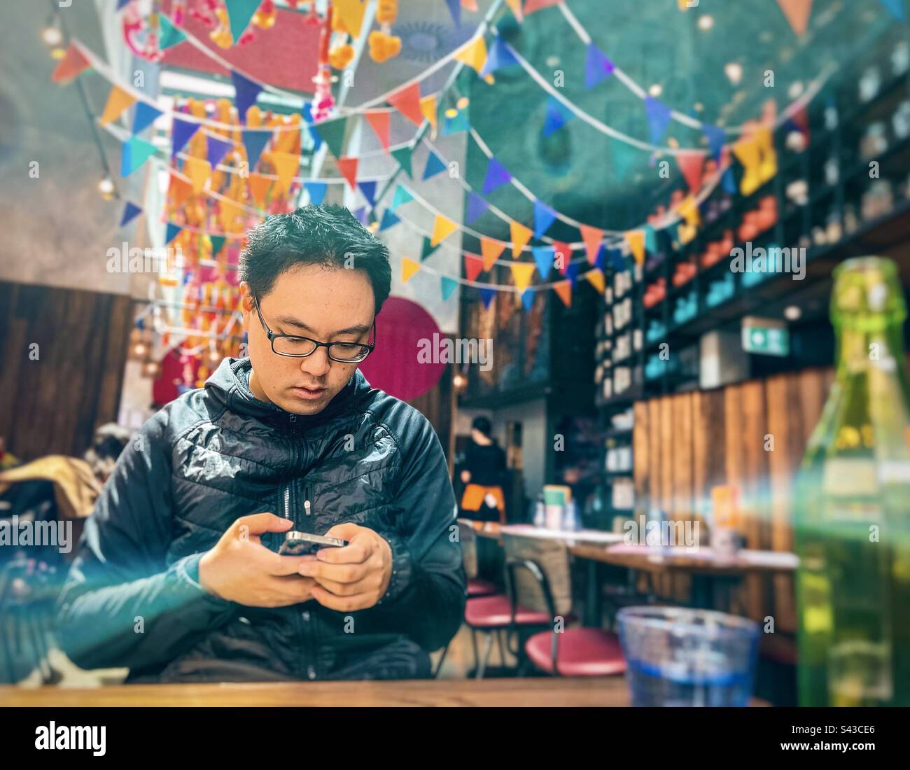 Jeune homme asiatique utilisant le téléphone portable à la table à l'intérieur du restaurant décoré avec des boutures colorées. - Image de stock capturée avec un smartphone