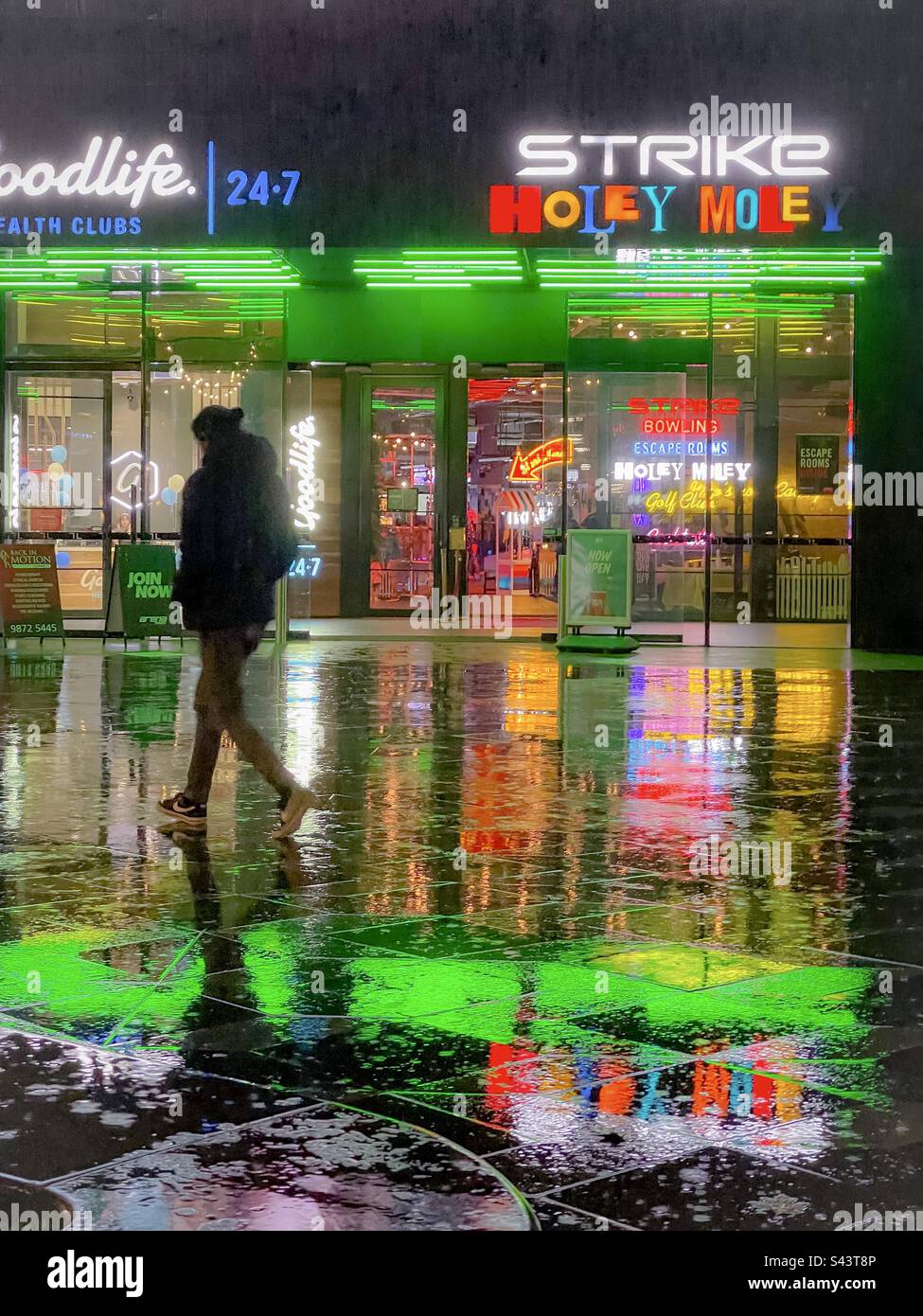 Scène nocturne pluvieuse, colorée et éclairée au néon, d'un homme qui marche sur des carreaux réfléchissants contre un centre d'amusement lumineux. - Image de stock capturée avec un smartphone