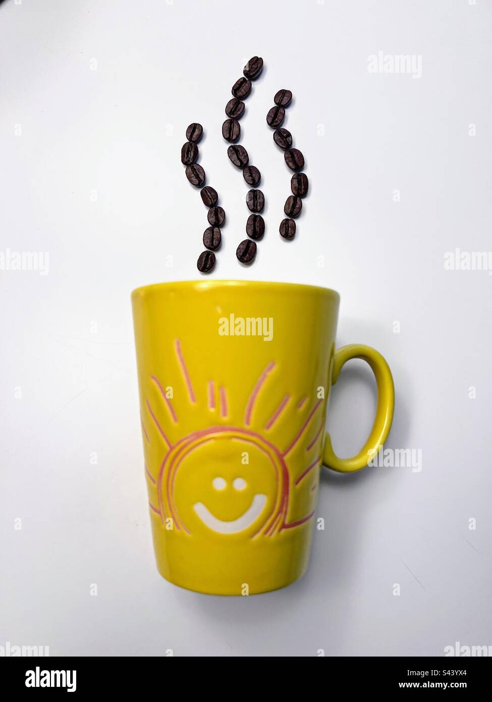 Image minimaliste abstraite avec tasse jaune avec dessin visage de soleil souriant et vapeur aromatique du matin représentée avec grains de café rôtis sur fond blanc - Image de stock capturée avec un smartphone