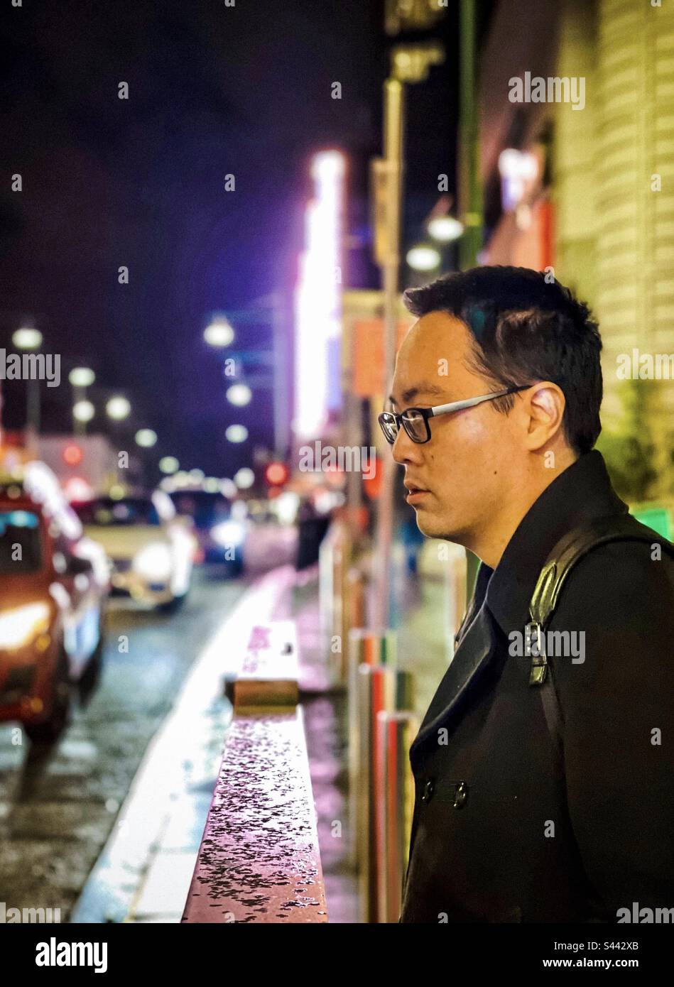 Portrait d'un jeune asiatique qui attend sur le trottoir contre les néons, les bâtiments et la route animée de la ville la nuit. Mise au point sélective. Coloré. - Image de stock capturée avec un smartphone