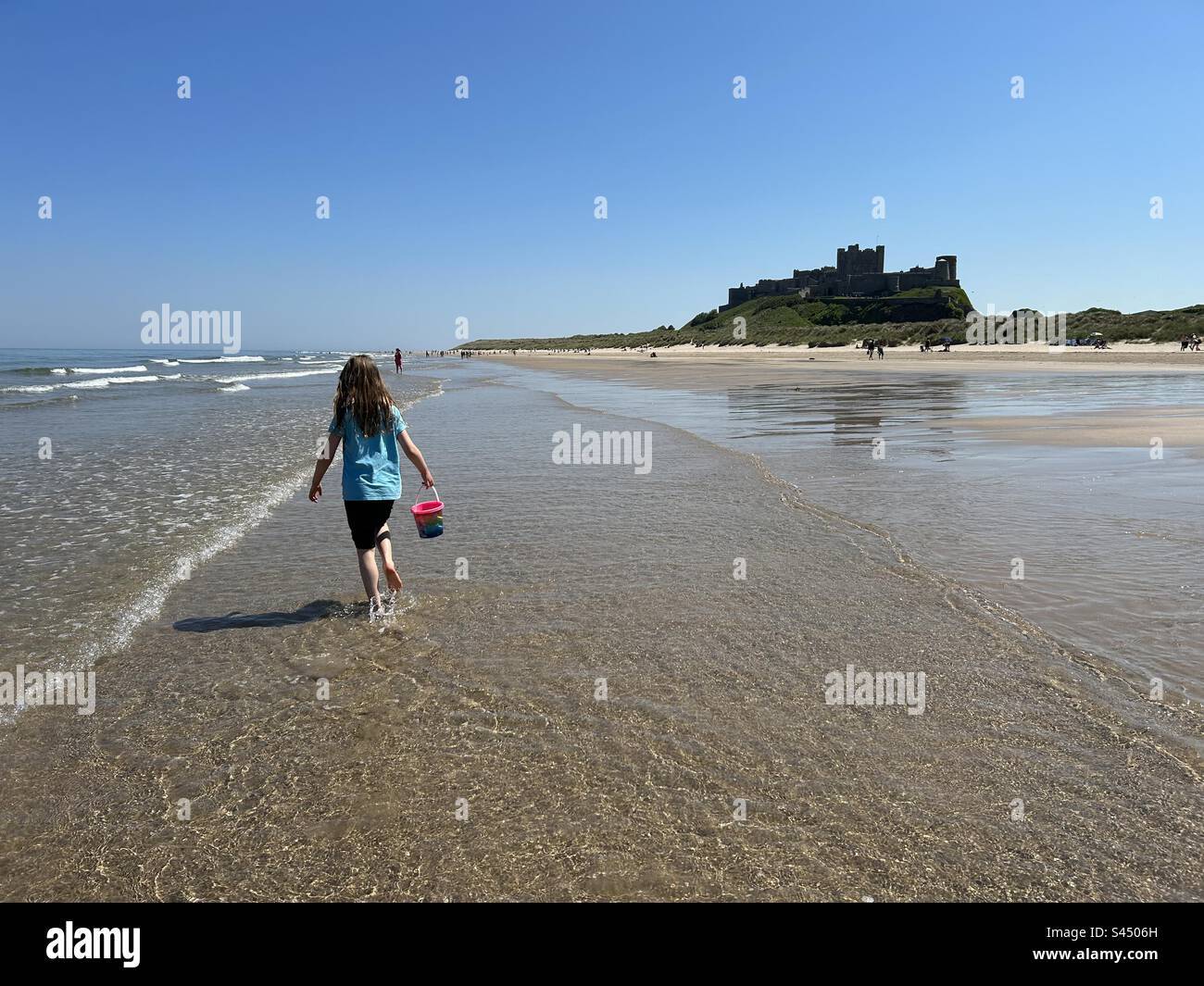 Fille marchant le long de la belle plage déserte de Bamburgh dans Northumberland tenant un seau rose, vue arrière. 8 ans Banque D'Images