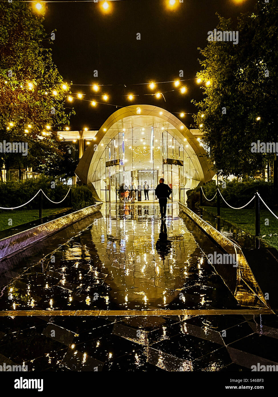 Un homme se précipitant dans l'entrée illuminée d'un centre commercial par une nuit humide et pluvieuse. Reflets, lumières de fées, nuit. Banque D'Images