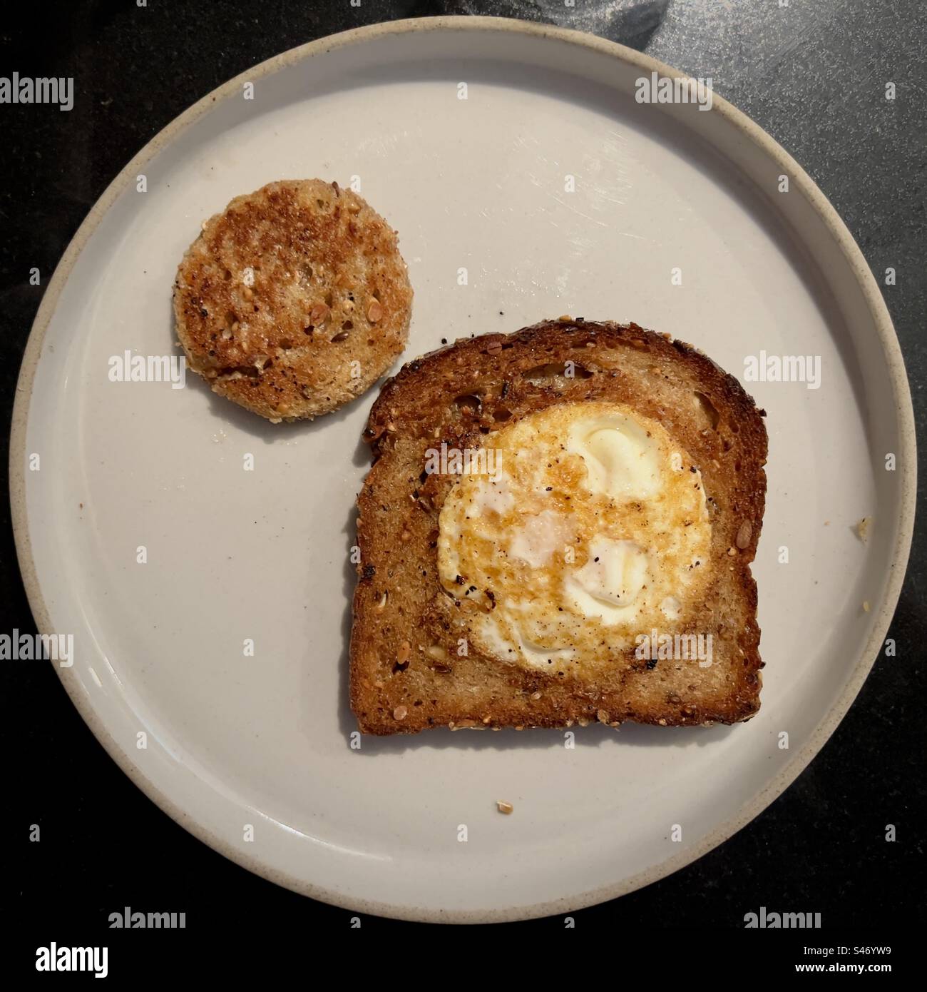 Oeuf frit dans un trou circulaire dans du pain frit, avec le trou découpé également frit, servi sur une assiette grise Banque D'Images Oeuf frit dans un trou circulaire dans du pain frit, avec le trou découpé également frit, servi sur une assiette grise Banque D'Images