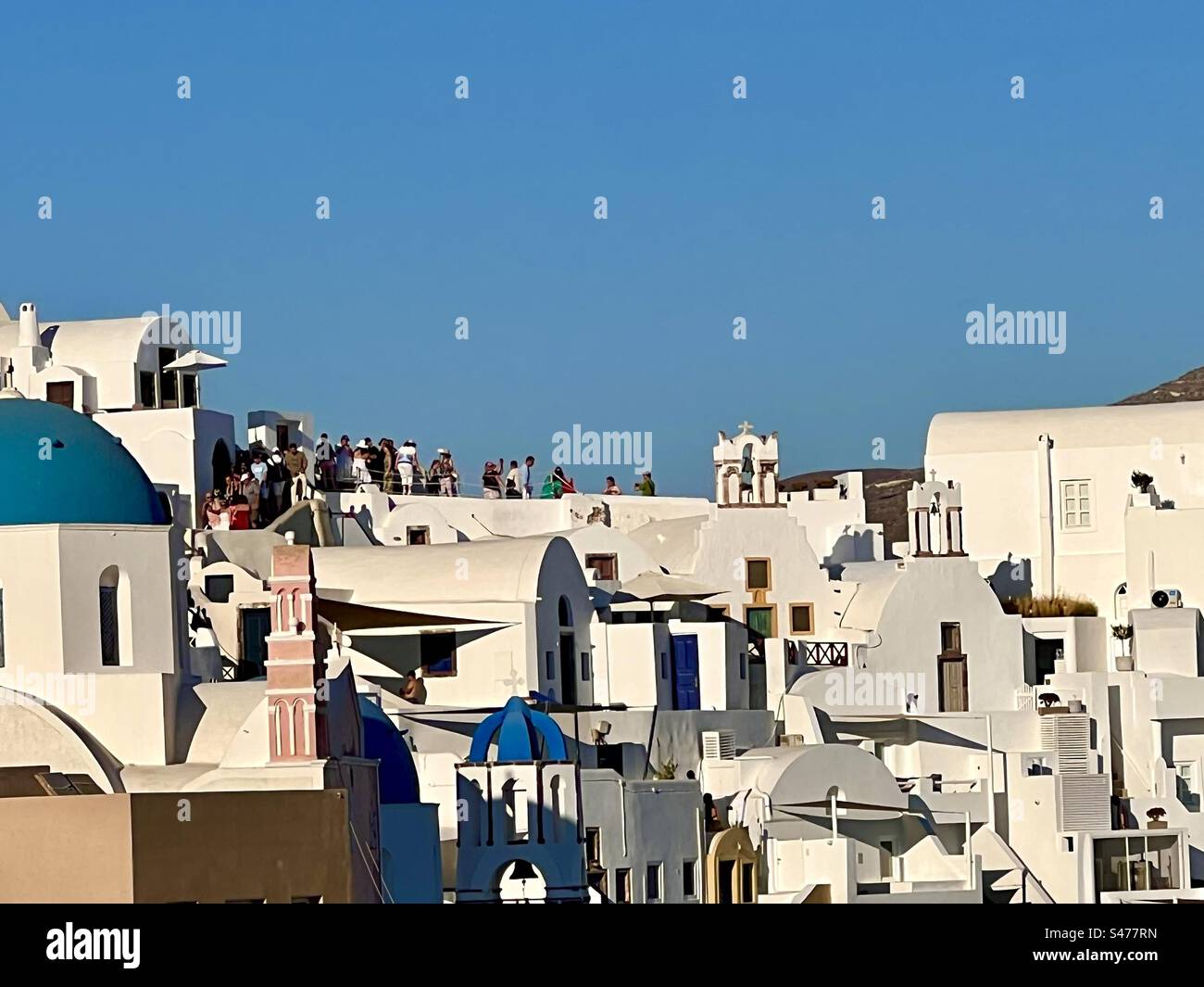 Vue sur les églises à dôme bleu et les bâtiments blanchis à la chaux de Santorin alors que les foules commencent à se rassembler dans les rues juste avant le coucher du soleil en été. - Image de stock capturée avec un smartphone