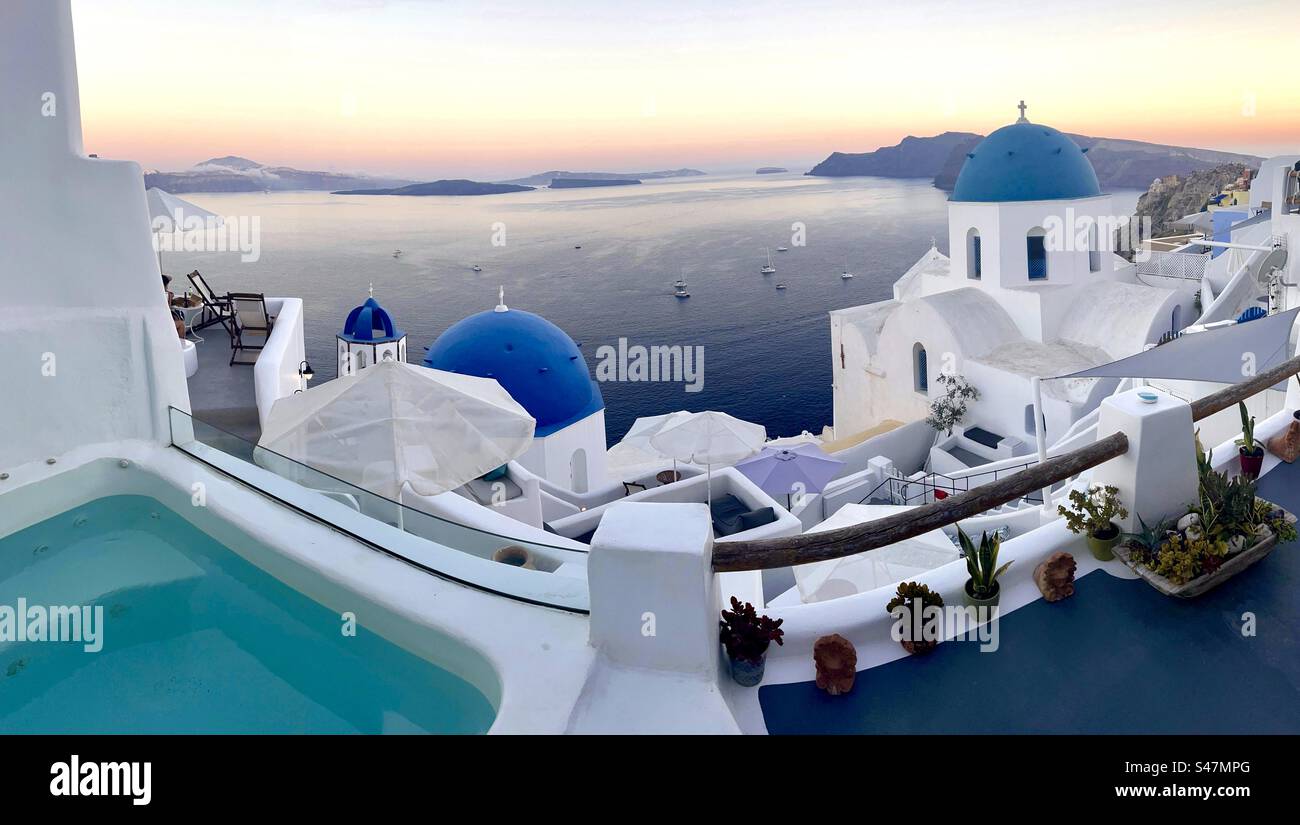 Vue panoramique depuis une terrasse perchée au-dessus de la caldeira à Oia, Santorin avec les tristement célèbres dômes bleus et horizon orange au coucher du soleil en été. - Image de stock capturée avec un smartphone