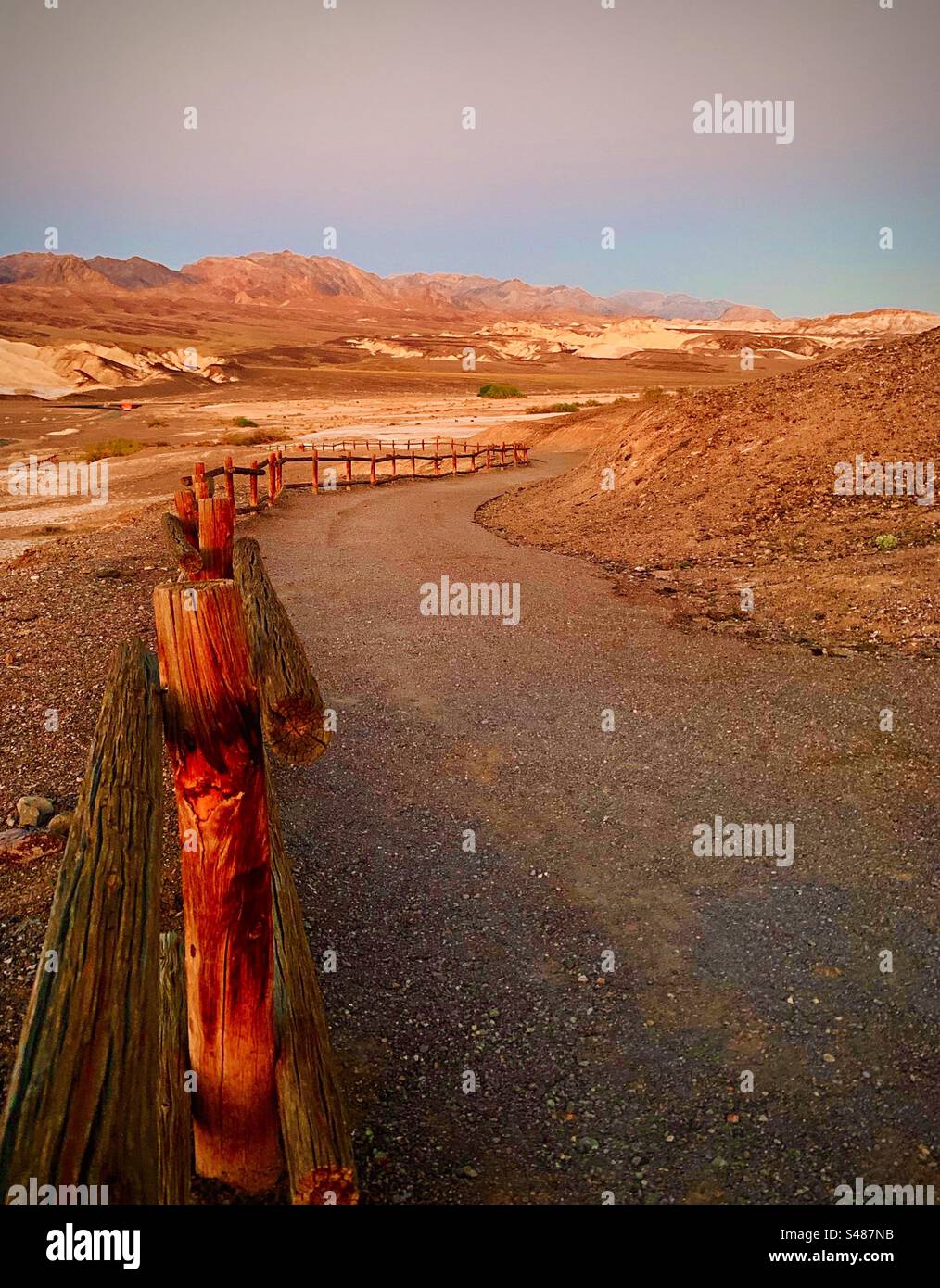 Une clôture s'étend le long d'un chemin dans un parc national de la Vallée de la mort en Californie, aux États-Unis. Il y a des rochers et du sable éparpillés autour du chemin de gravier et de hautes montagnes en arrière-plan. Ciel bleu. Banque D'Images