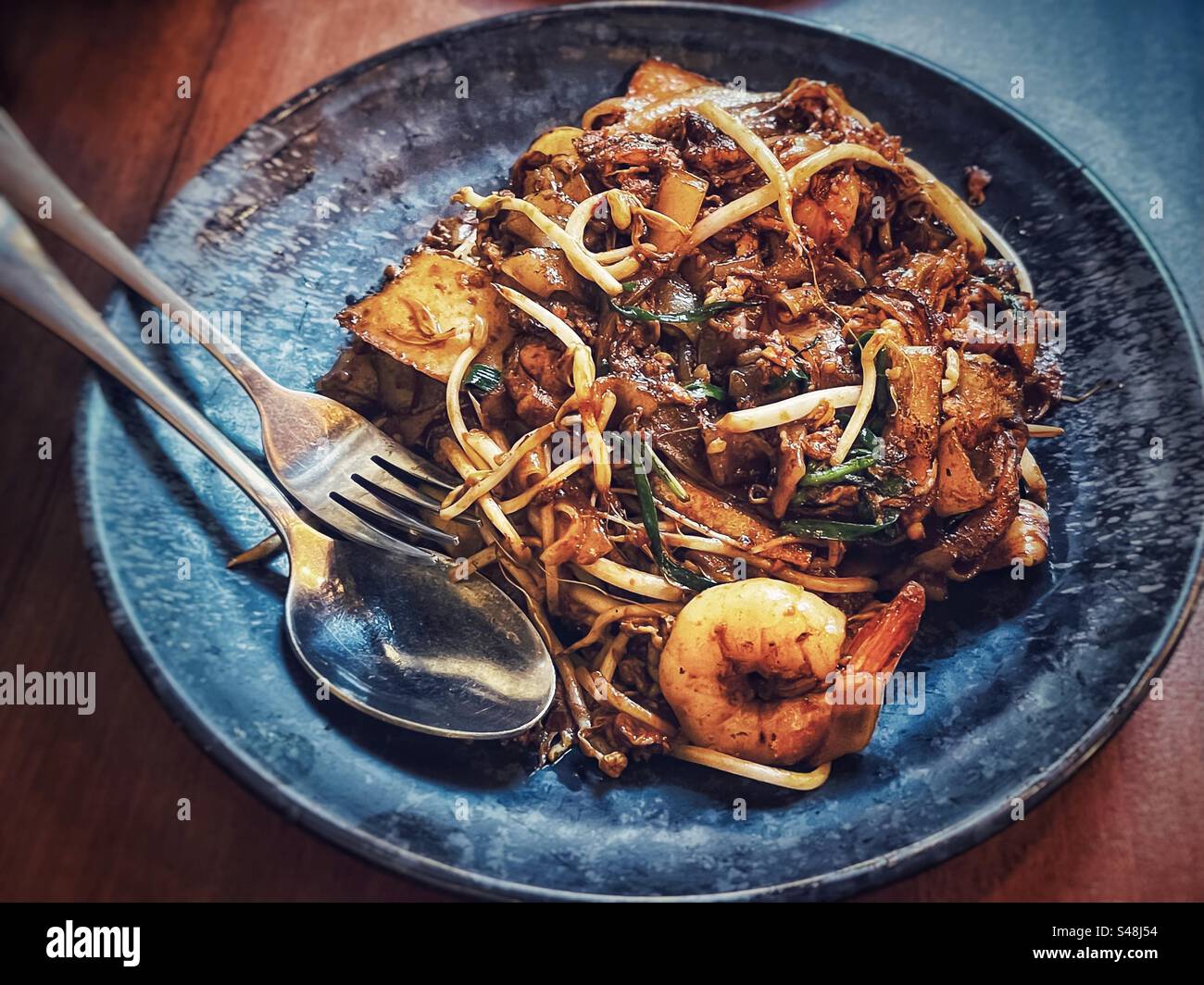 Vue à grand angle de char koay teow, nouilles plates frites épicées avec germes de haricot mungo, ciboulette et crevettes sur l'assiette avec fourchette et cuillère sur la table. Cuisine malaisienne. Banque D'Images