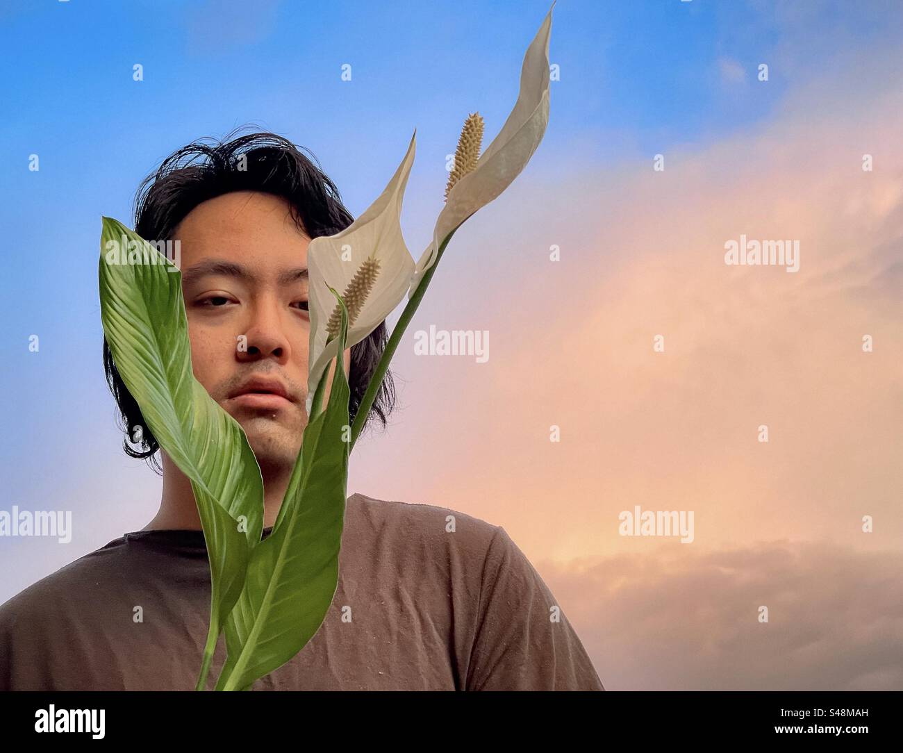 Portrait de jeune homme asiatique encadré par deux fleurs de lys de paix contre le ciel de coucher du soleil avec des nuages roses au printemps avec espace de copie. Banque D'Images