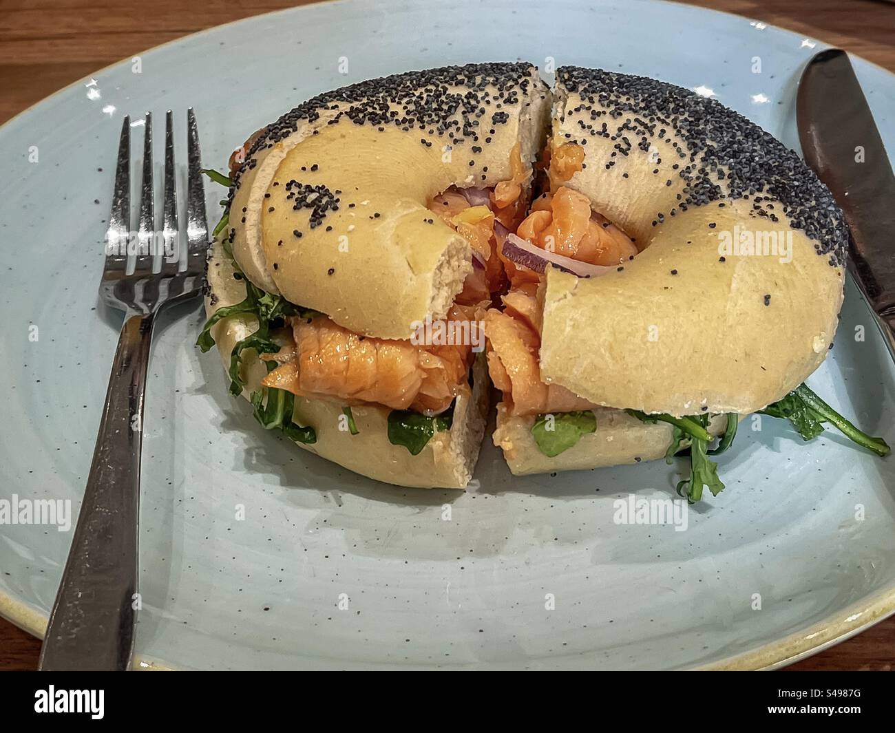 Gros plan de bagel de graines de pavot avec saumon fumé et légumes verts de salade sur une assiette avec couverts sur la table. - Image de stock capturée avec un smartphone