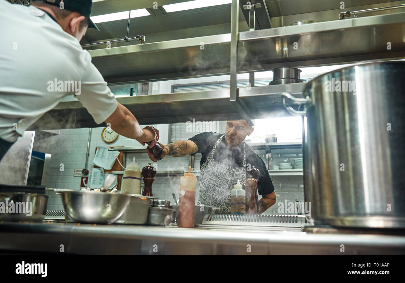 Prenez-le. Jeune chef professionnel en donnant l'aire d'une poivrière à son assistante, tout en processus de cuisson. Cuisine Restaurant Banque D'Images