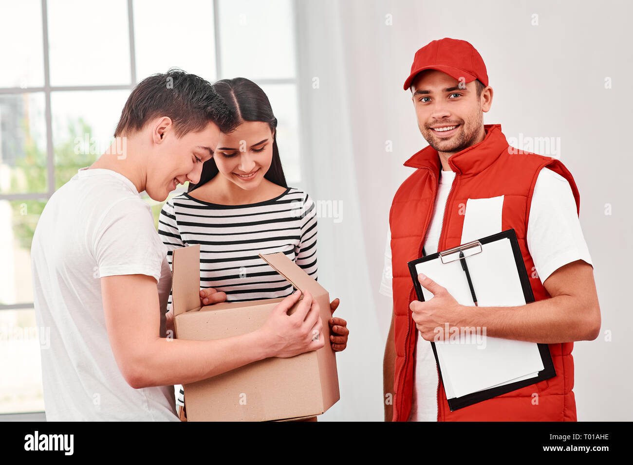 Cheerful postman porter du rouge uniforme postale est la livraison de colis à des clients satisfaits. Il apporte des boîtes en carton à son propriétaire. Il a fait son travail très bien. Dark-haired woman et colis ouvert de l'adolescence, ils sont heureux de le recevoir. Friendly travailleur, le service de haute qualité. À l'intérieur. Intérieur lumineux. Banque D'Images