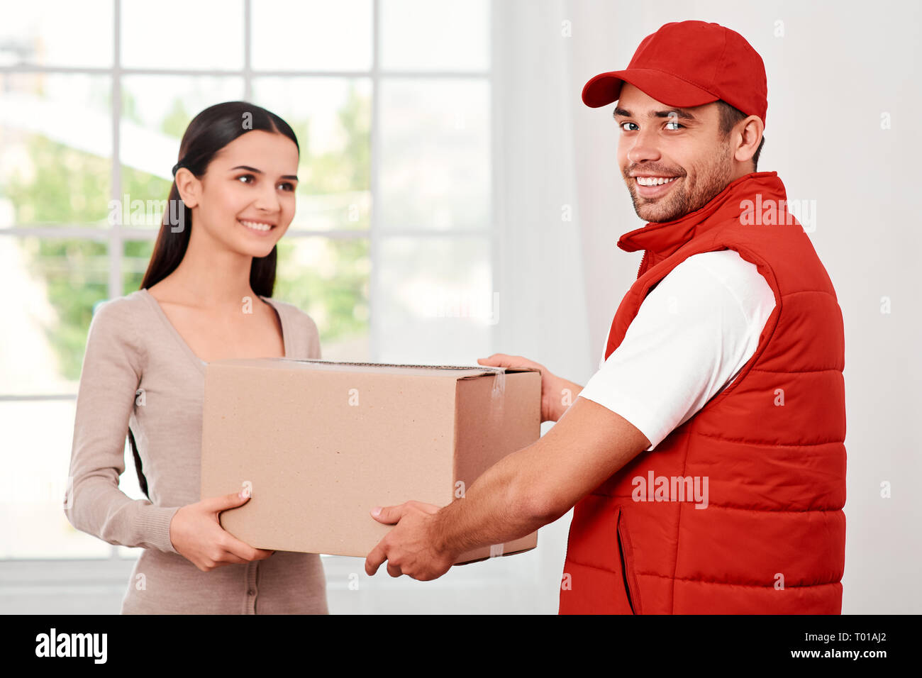 Cheerful postman porter du rouge uniforme postale est la livraison colis à un client satisfait. Il est en train de regarder l'appareil photo, alors qu'elle se penche sur lui. Ils détiennent en carton et souriant. Friendly travailleur, le service de haute qualité. À l'intérieur. Intérieur lumineux. Banque D'Images