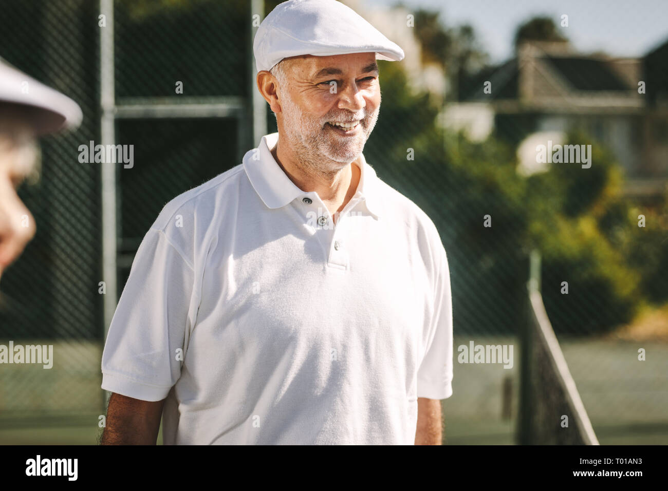 Cheerful senior homme debout sur un court de tennis sur une journée ensoleillée. Smiling man in white shirt de tennis et les standing outdoors. Banque D'Images