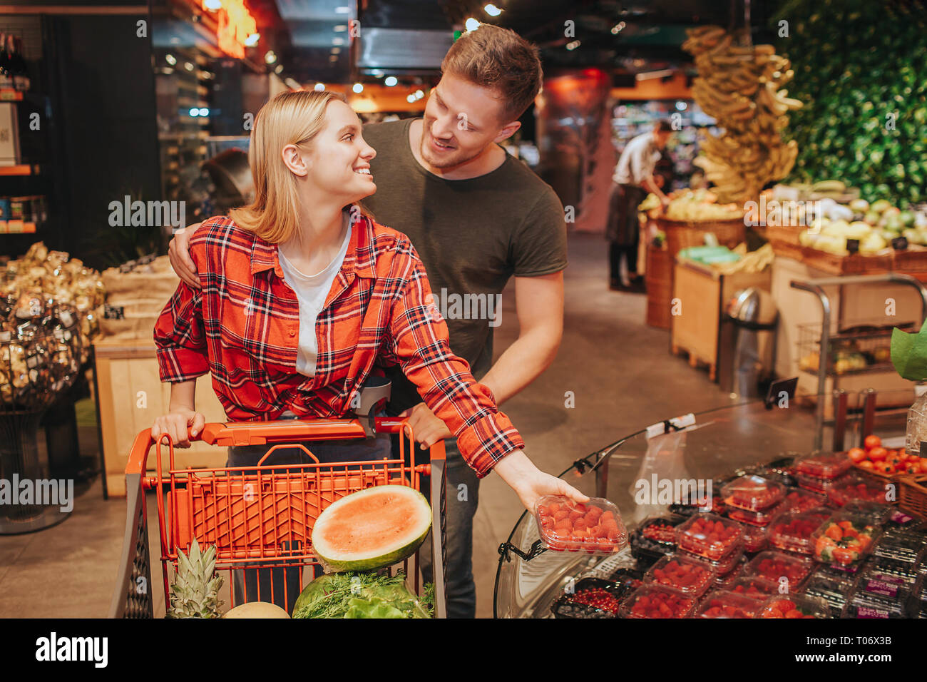 Jeune couple en épicerie. Elle se plaque avec de petits fruits et de regarder l'homme. Guy Se tenir derrière son chariot et le maintenir avec la main. Sourire à l'autre. Banque D'Images