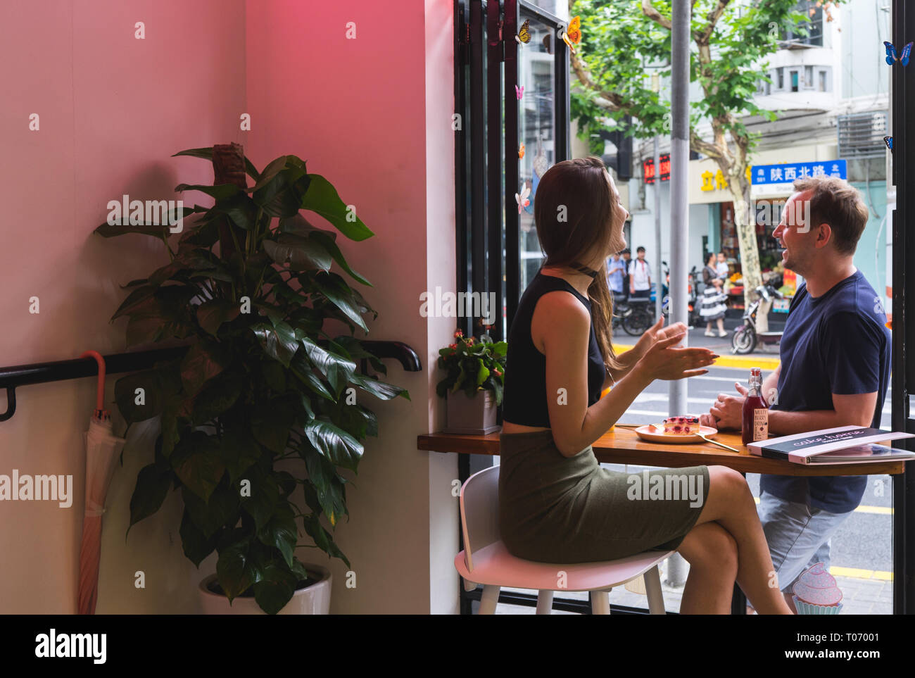 Un jeune couple caucasien, un homme et une femme, est assis dans un café et parle les uns avec les autres, fond rose, plante verte et rue à l'extérieur Banque D'Images