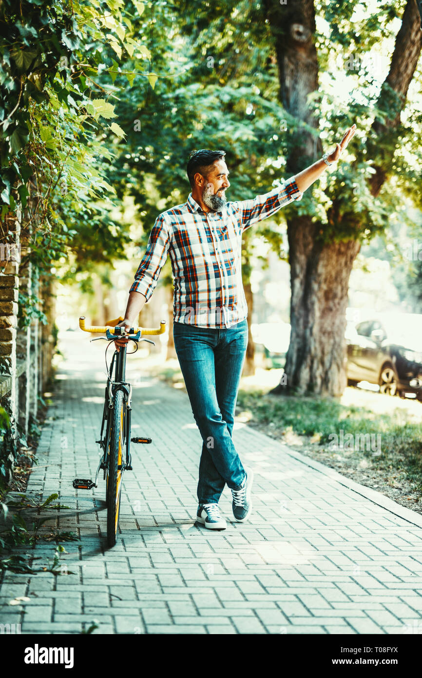 Un beau jeune homme va à la ville avec son vélo, debout à côté d'elle et en agitant à quelqu'un. Banque D'Images