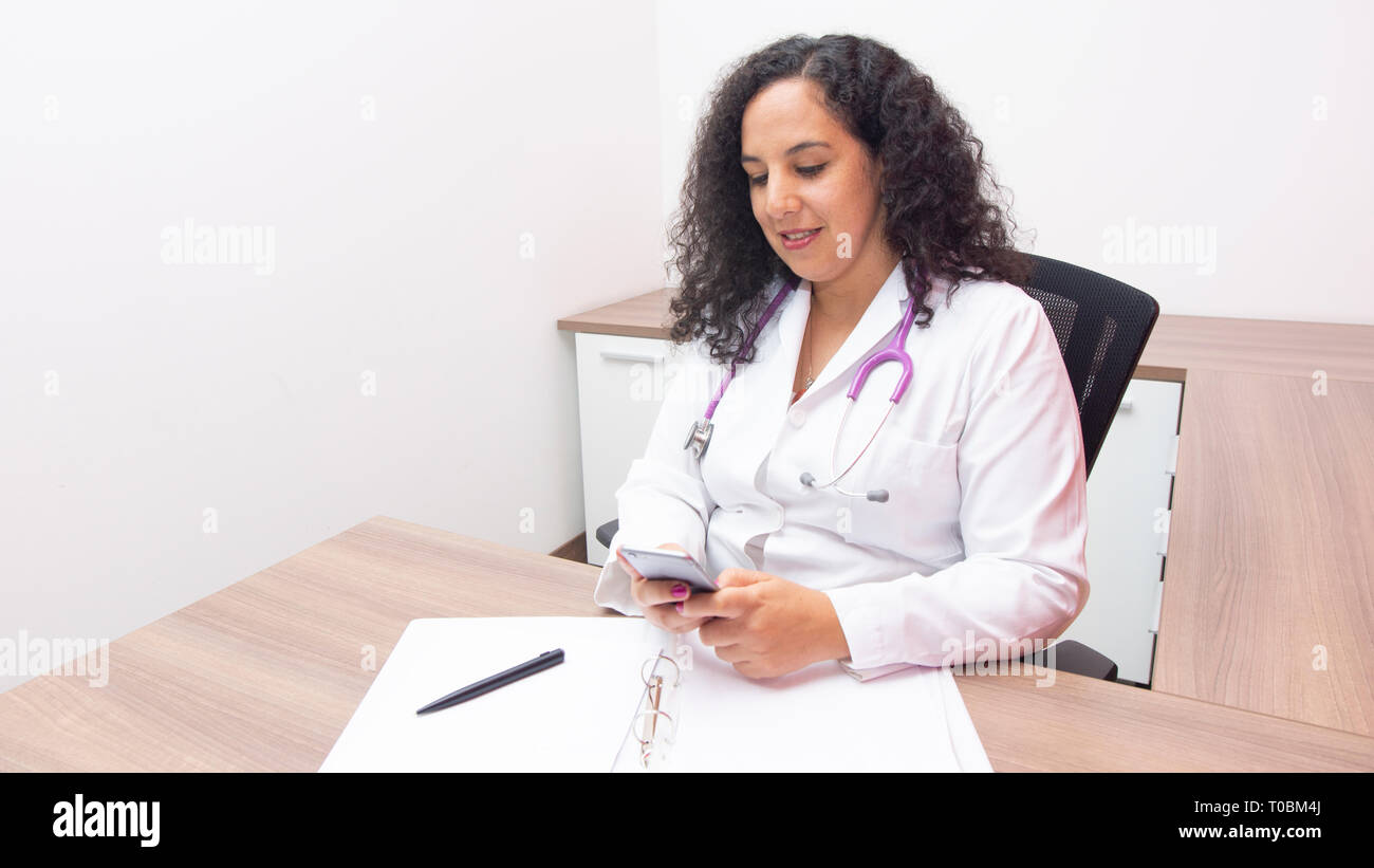 Femme médecin femme latine à la séance à son téléphone dans son bureau avec stéthoscope sur son cou de taper sur son smartphone Blackberry sur fond blanc Banque D'Images