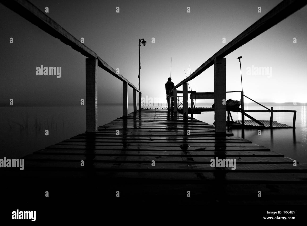 Silhouette d'un homme debout sur un quai en bois dans le noir, fixant le vide d'un lac avec feux lumineux sur le côté opposé Banque D'Images