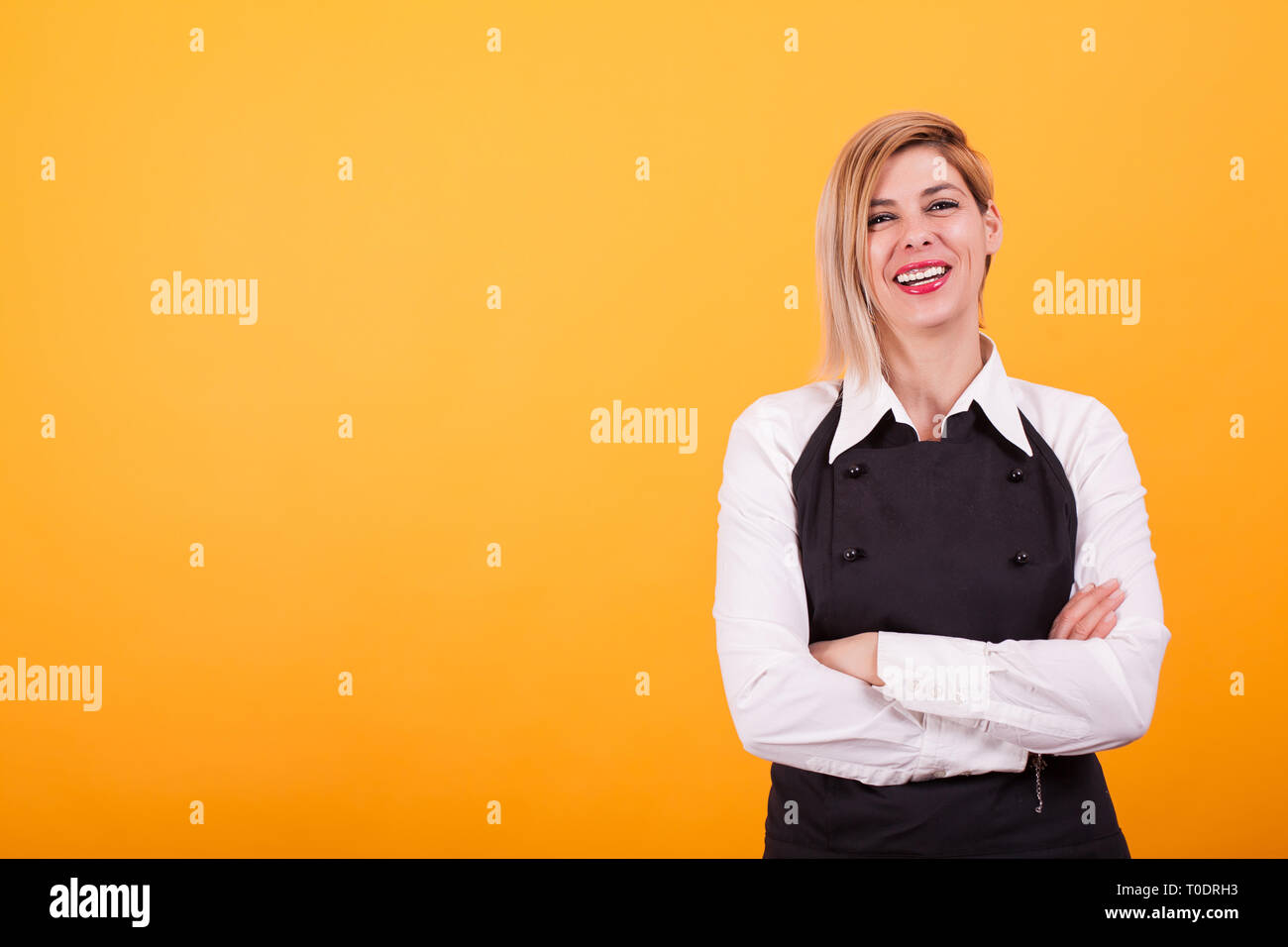 Très belle jeune femme blonde vêtue de son uniforme de Barman sur fond jaune. Bras croisés. T-shirt blanc. Banque D'Images