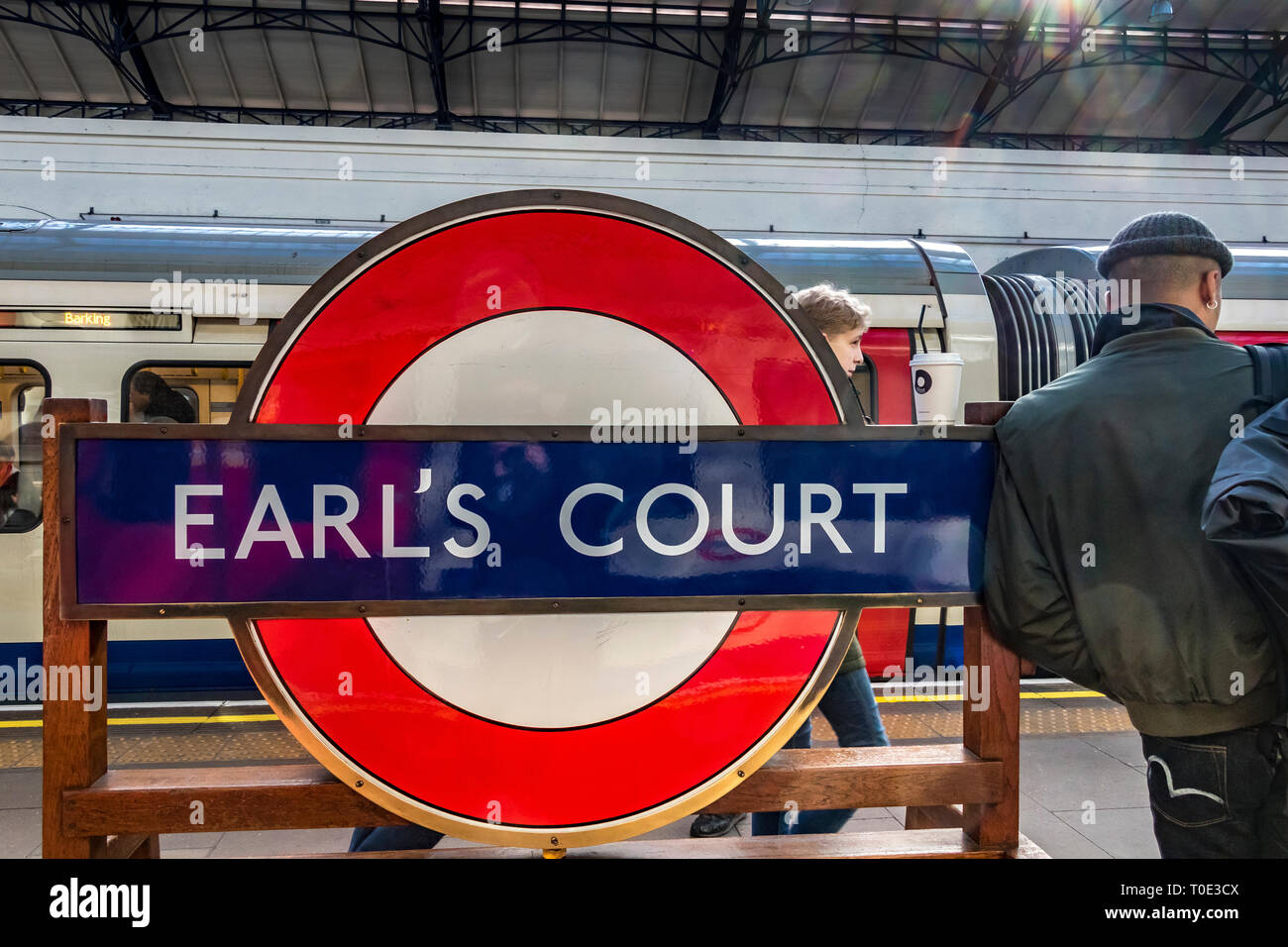 Un homme se penche contre le panneau rond de la station sur la plate-forme de la station de métro Earls court, Londres, Royaume-Uni Banque D'Images