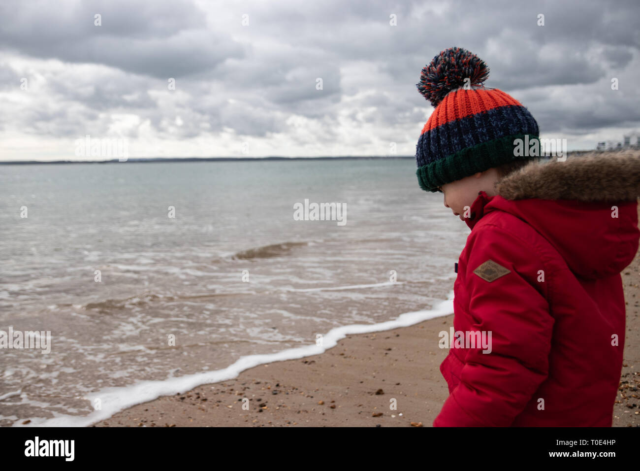 Jeune garçon de deux ans dans la région de Red Coat jouant sur la plage au printemps avec bobble hat Banque D'Images