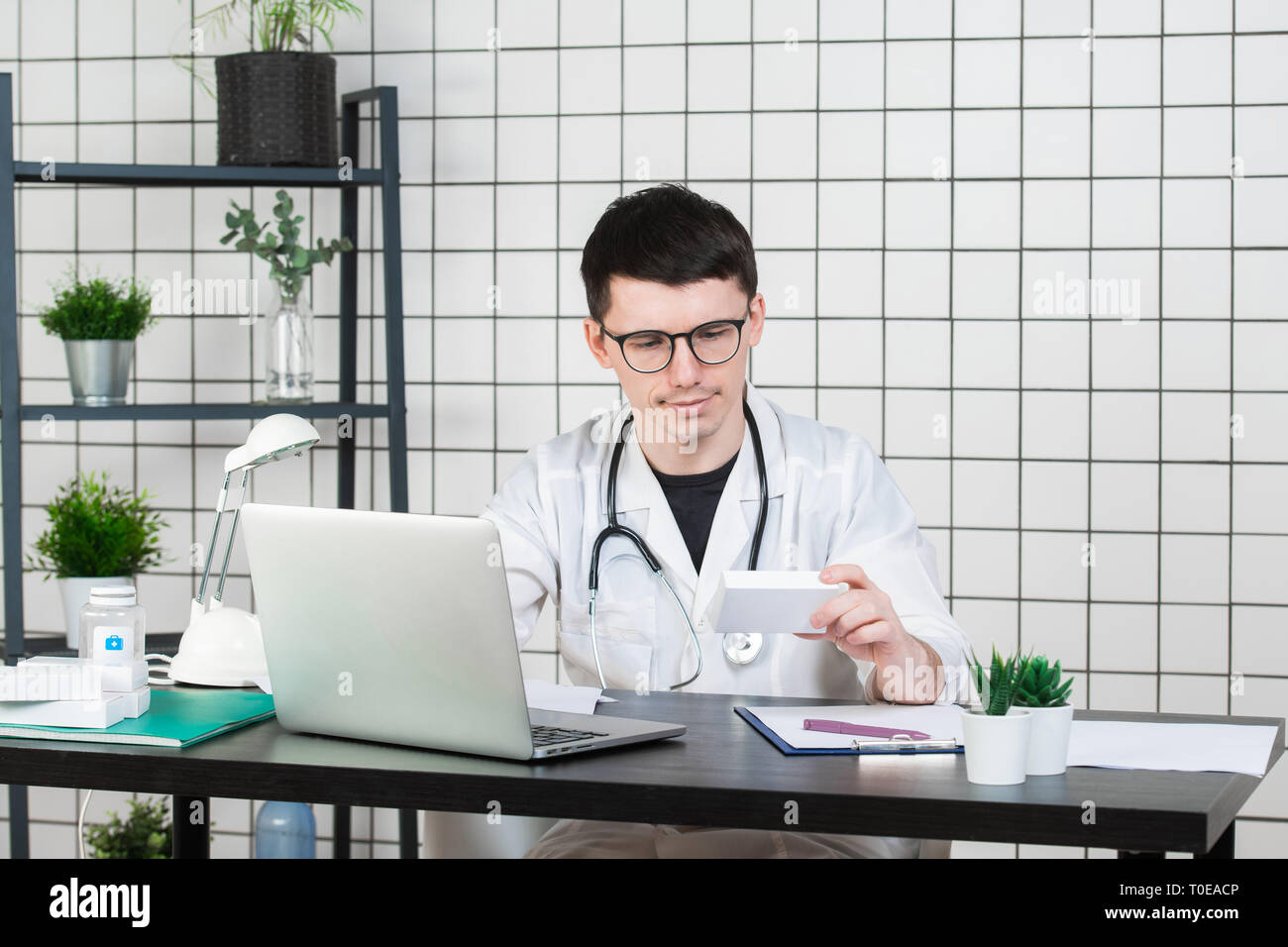 Médecin homme en blouse blanche avec stéthoscope sur son cou sitting at table pensant sur prescription, d'écrire quelque chose vers le bas, avec la boîte de médecine Banque D'Images