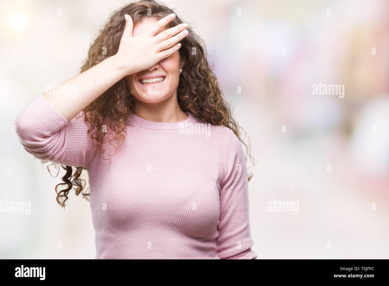 Belle brune cheveux bouclés young girl wearing sweater rose sur fond isolé sourire et rire avec la main sur le visage pour les yeux couvrant surpri Banque D'Images